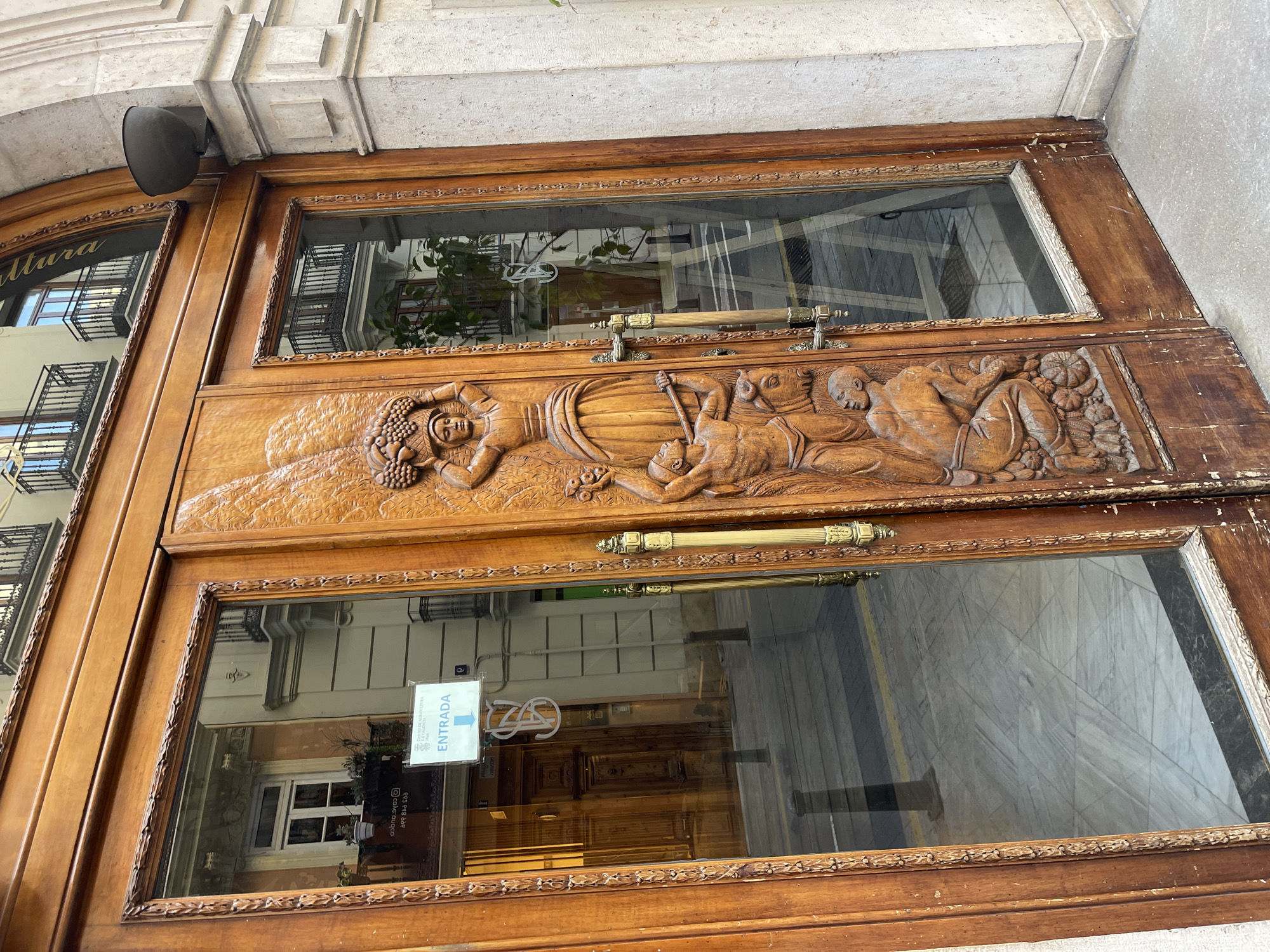 The image shows a set of ornate wooden doors with glass panels. A detailed wood carving depicting figures lies across the center of the doors. The carvings include a figure reclining with grapes, and other figures around them. Two golden handles span the width of the doors. The surrounding frame is also wood, ornately decorated. The doors likely belong to the building "Cobalto 15" on Carrer de les Barques 15, in València, Spain. In the reflection of the glass doors, the street and buildings across from the doors can be seen. A sign reading "ENTRADA" (Entrance) is attached to the lower glass panel. The door appears to be the entrance to the shop “Cobalto 15”.
