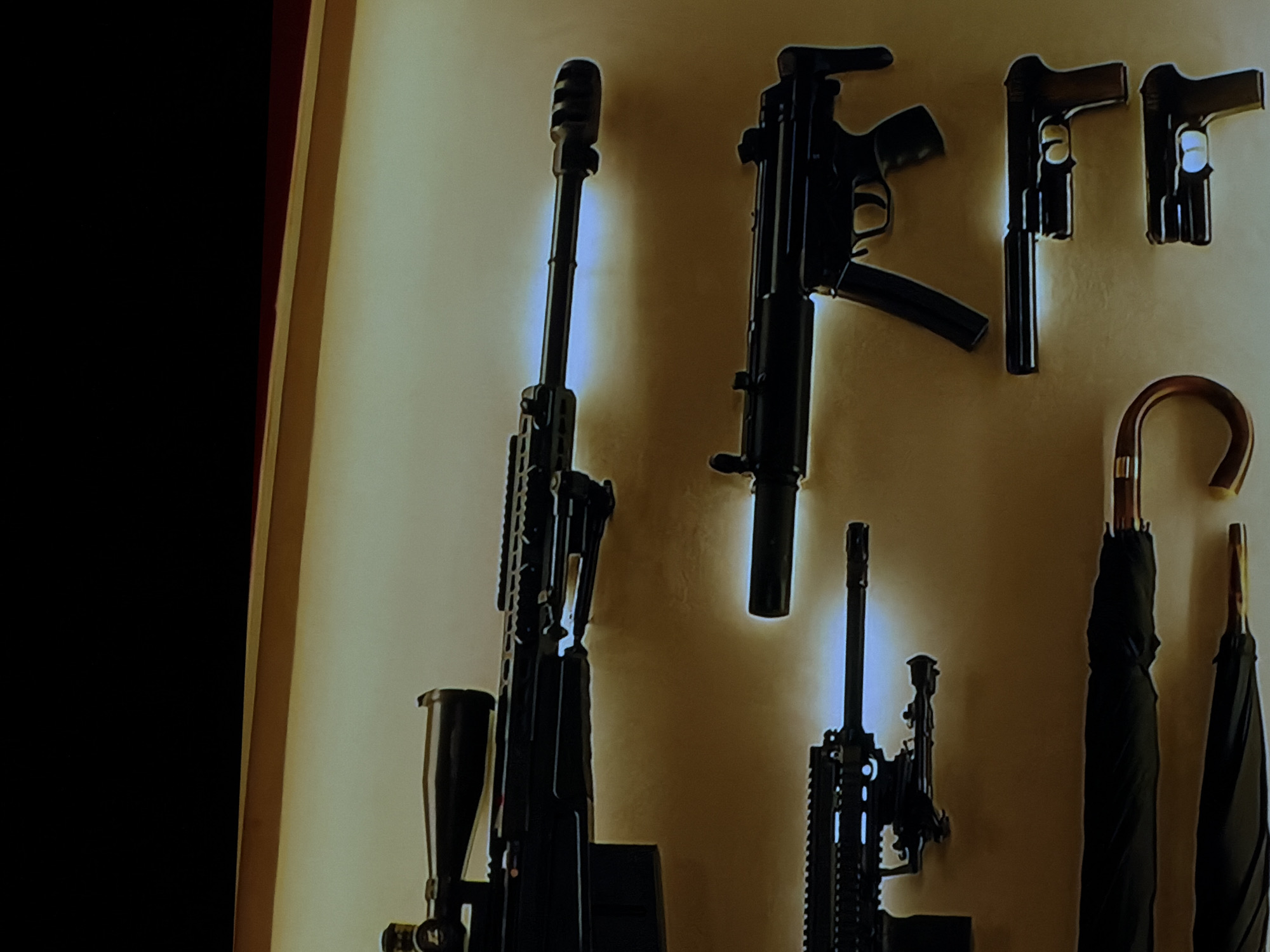 The image displays a collection of firearms and umbrellas mounted on a light-colored wall, likely indoors in a setting such as a private residence, armory, or collection display in Sofiivska Borshchahivka, Ukraine.
On the left side, a large, dark rifle, appearing to be a precision or sniper rifle, is positioned vertically. It features a prominent muzzle brake, a long barrel, and a scope mounted above the receiver. To its right and slightly above, a submachine gun, resembling an MP5 variant, is also mounted vertically. This firearm has a magazine extending downwards and a stock visible at the top. Below and slightly to the right of the submachine gun, another rifle, possibly an assault rifle or carbine, is partially visible, also mounted vertically.
In the upper right section of the wall, two dark handguns (pistols) are displayed horizontally. The one on the left appears to have a suppressor attached to its barrel. The handgun to its right has a visible light-colored grip panel.
On the far right side of the image, two closed black umbrellas with distinctive curved wooden handles are hanging vertically. The lighting in the scene creates subtle highlights around some of the displayed items, suggesting they might be illuminated by an overhead light source. A dark area on the far left of the frame indicates a boundary, possibly a doorway or another wall. No specific activity is taking place, and no text is visible within the image.
