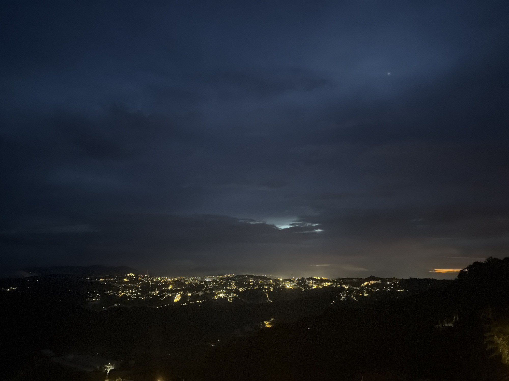 This is a nighttime aerial view of a city illuminated by artificial lights. The city is nestled within rolling hills, with the lights forming a dense cluster across the landscape. A dark, cloudy sky looms above, with a single bright star or planet visible in the upper right quadrant. The foreground is dominated by dark, silhouetted hills and foliage. The city lights suggest a densely populated area, possibly a town or small city, with buildings and streets clearly outlined by the warm glow. The overall atmosphere is serene and slightly mysterious, with the darkness of the night contrasting sharply with the vibrant urban lights.
Based on the provided context, the location is Xuân Trường, Đà Lạt, Vietnam. The image depicts the city of Xuân Trường at night, characterized by its widespread illumination from residential and commercial buildings, spreading across the undulating terrain typical of Đà Lạt's landscape. The silhouette of surrounding hills and vegetation is prominent in the foreground and along the city's periphery. The sky exhibits a dark, overcast condition, with a notable celestial point of light high up.