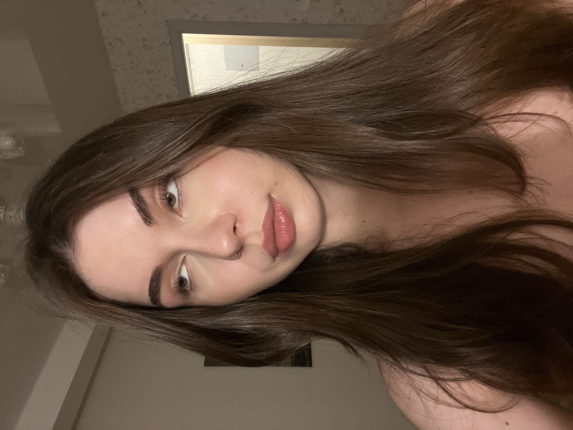 A young woman with long brown hair is photographed from an upward angle. Her head is tilted, and she looks at the camera. She has fair skin, defined eyebrows, and full lips. The background includes a blurry room interior with a glimpse of a doorway. The location, Saint Petersburg, Russia, cannot be determined from the visual information provided in the image.