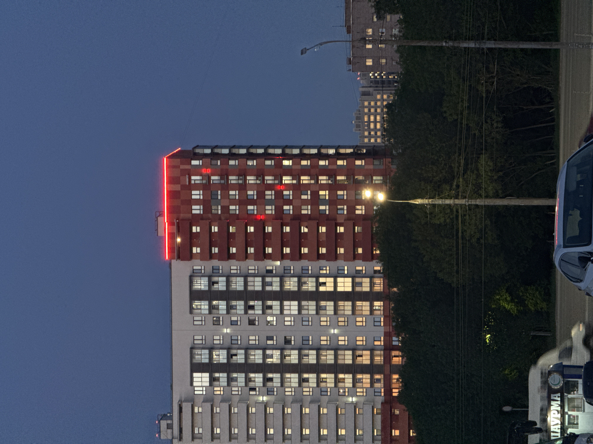 The image depicts a multi-story residential building in Izhevsk, Russia, at dusk. The building is divided into a lower section of white with dark gray accents and an upper section of red brick. A red neon light strip illuminates the top edge of the building. Many windows are lit from within. To the right of the building is a row of dense trees, a street, and several parked cars. A street lamp is situated in front of the trees. Another similar building is visible in the background to the upper left. The sky is a deep blue. The image is oriented vertically.