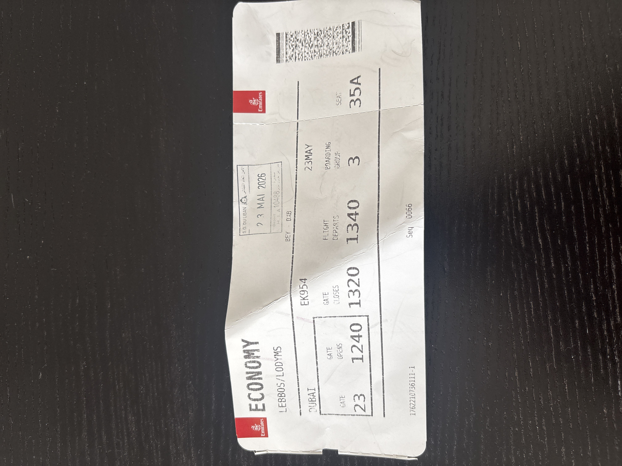A crumpled airline ticket is laid out on a dark wood textured surface. The ticket is predominantly white with red and black text and logos. It is an economy class ticket for Emirates airline. The ticket indicates a departure from Dubai, United Arab Emirates, to an unknown destination. Key details visible include:
* **Airline:** Emirates
* **Class:** ECONOMY
* **Origin:** DUBAI
* **Flight Number:** EK954
* **Date:** 23 MAY 2026
* **Gate:** 23
* **Gate Opens:** 1240
* **Gate Closes:** 1320
* **Flight Departs:** 1340
* **Boarding Group:** 3
* **Seat:** 35A
The surface appears to be a table with visible wood grain. The lighting suggests an indoor setting, with the ticket casting a slight shadow. There are no people or other objects visible in the image.