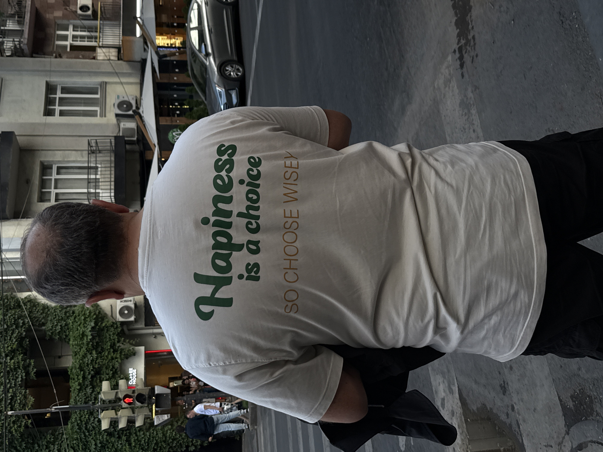The image captures an urban street scene in Yerevan, Armenia, from a slightly elevated perspective, focusing on the back of a person wearing a white t-shirt. The main subject is seen from the shoulders down, with a close-up of their head and upper back, where the t-shirt prominently displays text. The text, in green, reads "Happiness is a choice," with "SO CHOOSE WISELY" in a smaller, brownish font underneath.
The person appears to be walking or standing on a paved street or sidewalk. To their left, there are multi-story buildings with windows and balconies, some adorned with lush green ivy or similar climbing plants. Several air conditioning units are visible on the building facades, one partially showing the brand "BOSCH." Further down the street, other pedestrians are visible, though blurred. A traffic light showing a red signal is also in the foreground on the left.
To the right of the main subject, a dark sedan is parked or driving on the street. The ground is a mix of dark pavement and lighter concrete. The lighting suggests it is daytime, possibly late afternoon, with clear weather. Another partial sign on a building in the background reads "CHA E HOUSE." The overall setting depicts a busy city environment with both residential or commercial buildings and street activity.
