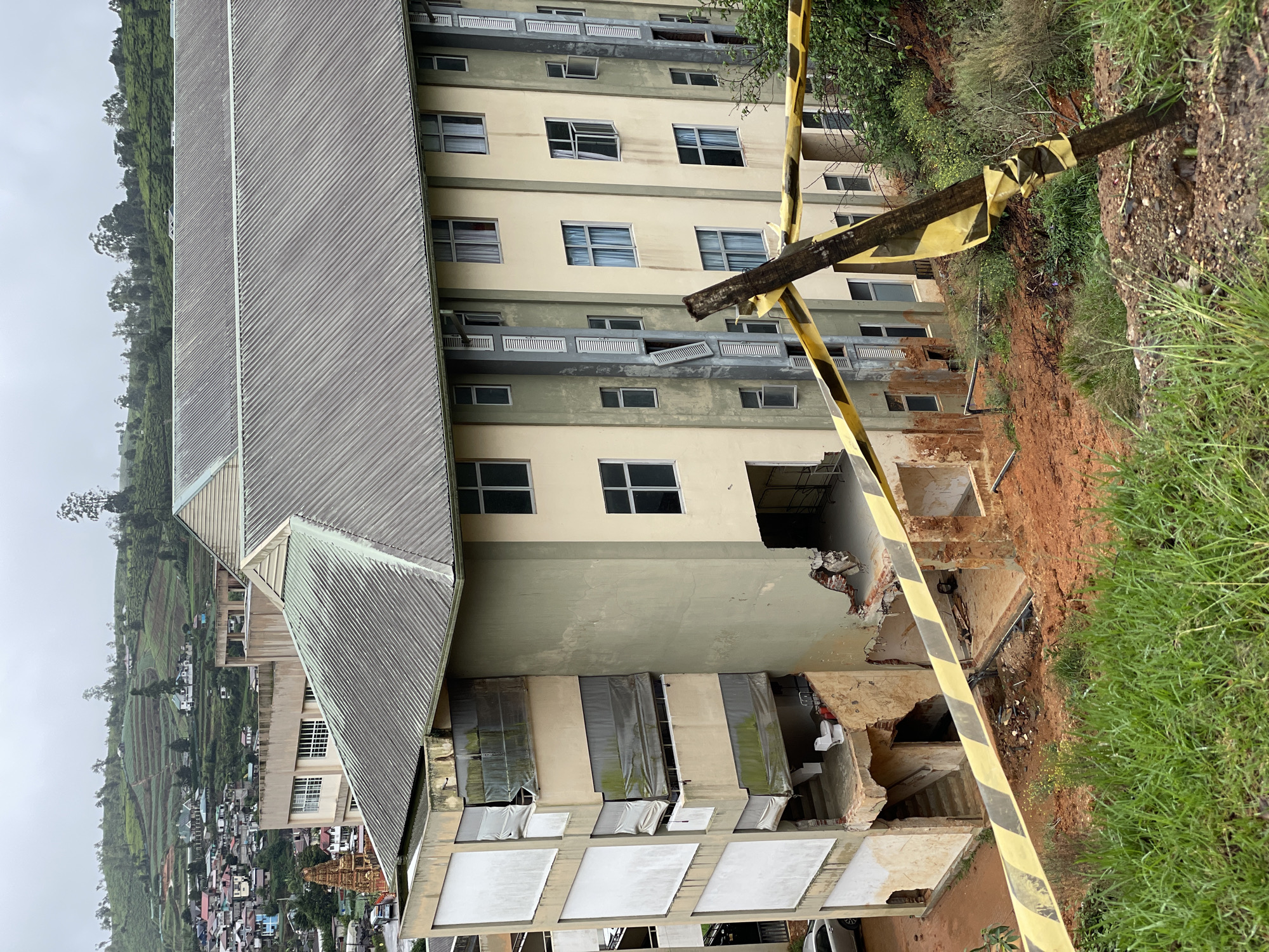 A building in Nuwara Eliya, Sri Lanka, shows significant damage, likely from a landslide, with exterior walls collapsed and portions of the structure exposed. Yellow and black caution tape surrounds the affected area. The building is multi-story with a pitched roof. The surrounding environment includes greenery, soil, and what appears to be urban development in the background.