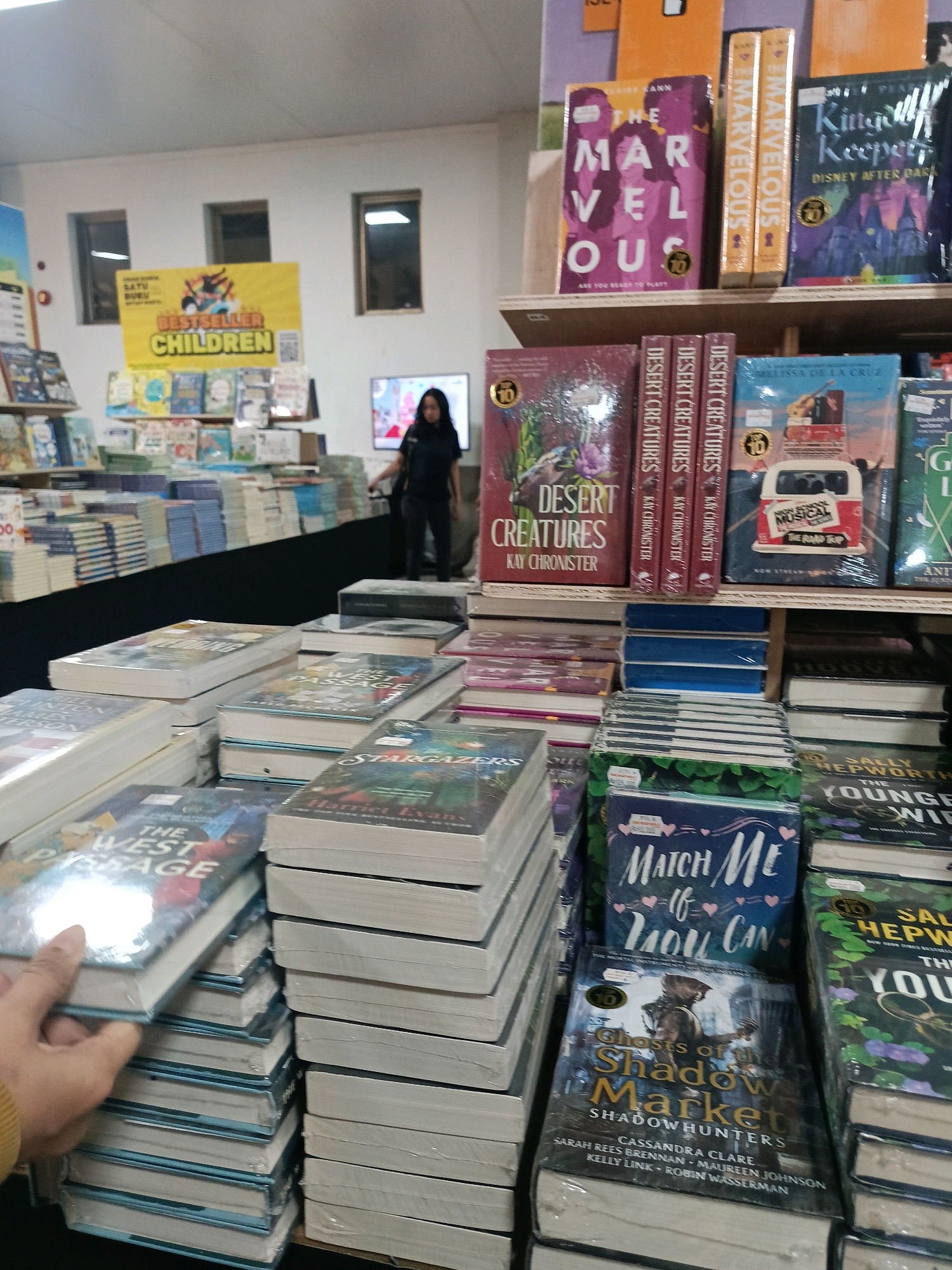 The image shows an indoor setting, likely a bookstore, filled with shelves of books. In the foreground, a hand is reaching out to pick up a book titled "THE WEST PASSAGE." Various books are stacked neatly on tables and shelves, with titles such as "DESERT CREATURES," "MARVELOUS," "KITTEN KEEPER," "HIGH SCHOOL MUSICAL THE ROAD TRIP," "MATCH ME IF YOU CAN," and "GHOSTS OF THE SHADOW MARKET." In the background, a woman wearing a dark top and pants is standing near a television screen. A yellow banner with "BESTSELLER CHILDREN" is visible on a shelf. The overall environment suggests a commercial establishment selling books. The presence of the "Satu Buku" logo on the yellow banner indicates the location is likely in Banguntapan, Indonesia, as "Satu Buku" is a known bookstore chain in Indonesia.