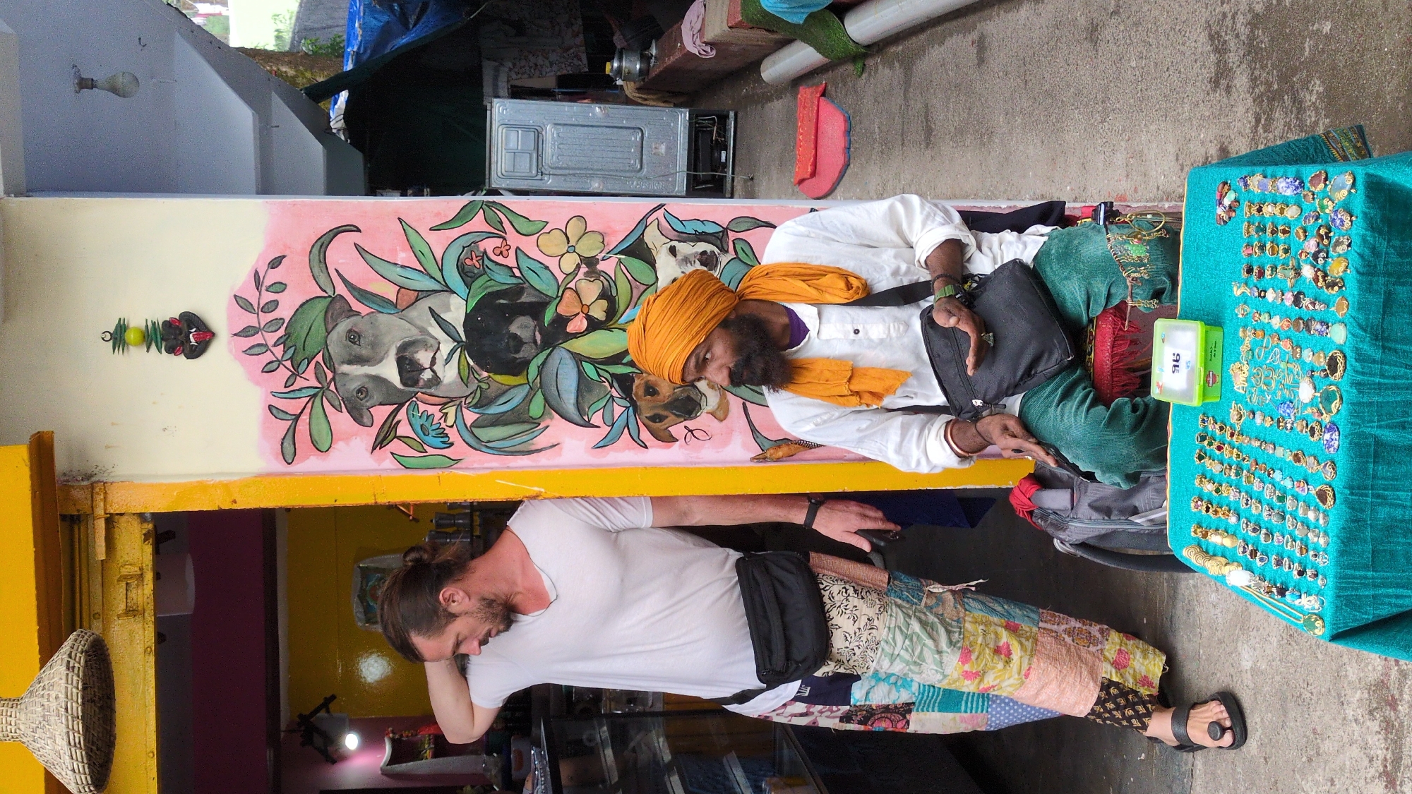 This image captures a vibrant street scene in Bhagsu, India, featuring two men and an array of merchandise. The setting appears to be an outdoor market or a shop's entrance, characterized by a colorful mural on the wall.
On the right, a man with dark skin, a thick black beard, and an orange turban is seated, wearing a white shirt and green pants. He holds a black bag on his lap and looks towards a table covered in a teal cloth, which displays numerous pieces of jewelry, possibly earrings or pendants, arranged in rows. A small, clear green plastic container with a lid sits on the table, revealing faint white text that includes "Rs." and "95," likely indicating a price. This man appears to be a vendor, observing his goods.
To the left, another man with lighter skin, a beard, and his hair tied in a bun stands, leaning against a yellow structure that frames a shop entrance. He wears a white t-shirt, a black fanny pack slung across his chest, and distinctive multi-colored patchwork pants. He looks downwards, seemingly contemplative or browsing.
The wall behind the seated man features a prominent mural with artistic depictions of dog faces, possibly Pit Bulls, intertwined with green foliage and light pink and blue floral elements, set against a background that transitions from light pink to cream. Above the standing man, a large woven basket, possibly a lampshade, hangs. The ground is concrete. The overall lighting suggests daytime. The visible text "Rs. 95" is on the green container.