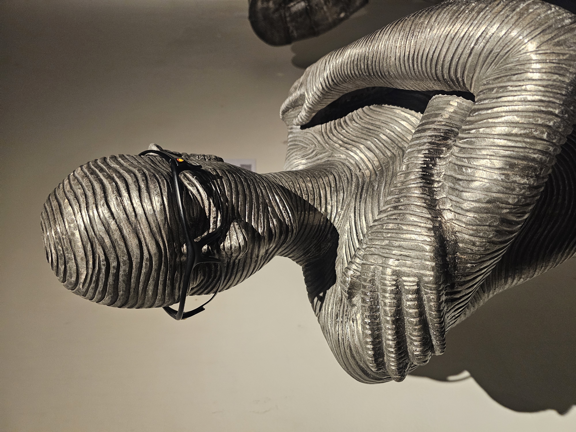 A sculpture of a human figure constructed from horizontally ridged metal is captured in a close-up shot. The figure is posed with the head bowed and hands clasped near the lower body. A pair of glasses rests on the head of the statue. The sculpture is situated inside the Griya Santrian a Beach Resort in Denpasar, Indonesia.