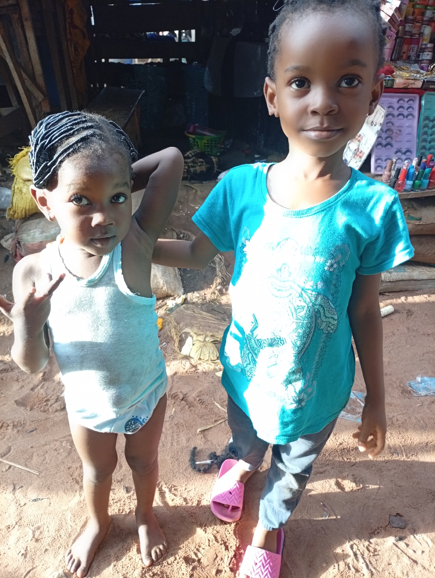 Two young Black girls stand on a sandy surface in what appears to be an outdoor marketplace or informal settlement in Nkpor, Nigeria. The girl on the left, wearing a white tank top and light blue underwear with a cartoon character on it, has her hair styled in thin braids with black beads. She is looking towards the camera with a curious expression, her left hand raised. The girl on the right, wearing a bright teal t-shirt with a white patterned design and dark pants, gazes directly at the camera with a neutral expression. She is wearing pink slides. Behind the girls, there are wooden structures, possibly market stalls, and various objects like sacks, netting, and colorful items that could be merchandise. The lighting suggests it is daytime, with strong sunlight casting shadows. The ground is covered in sand and some debris.