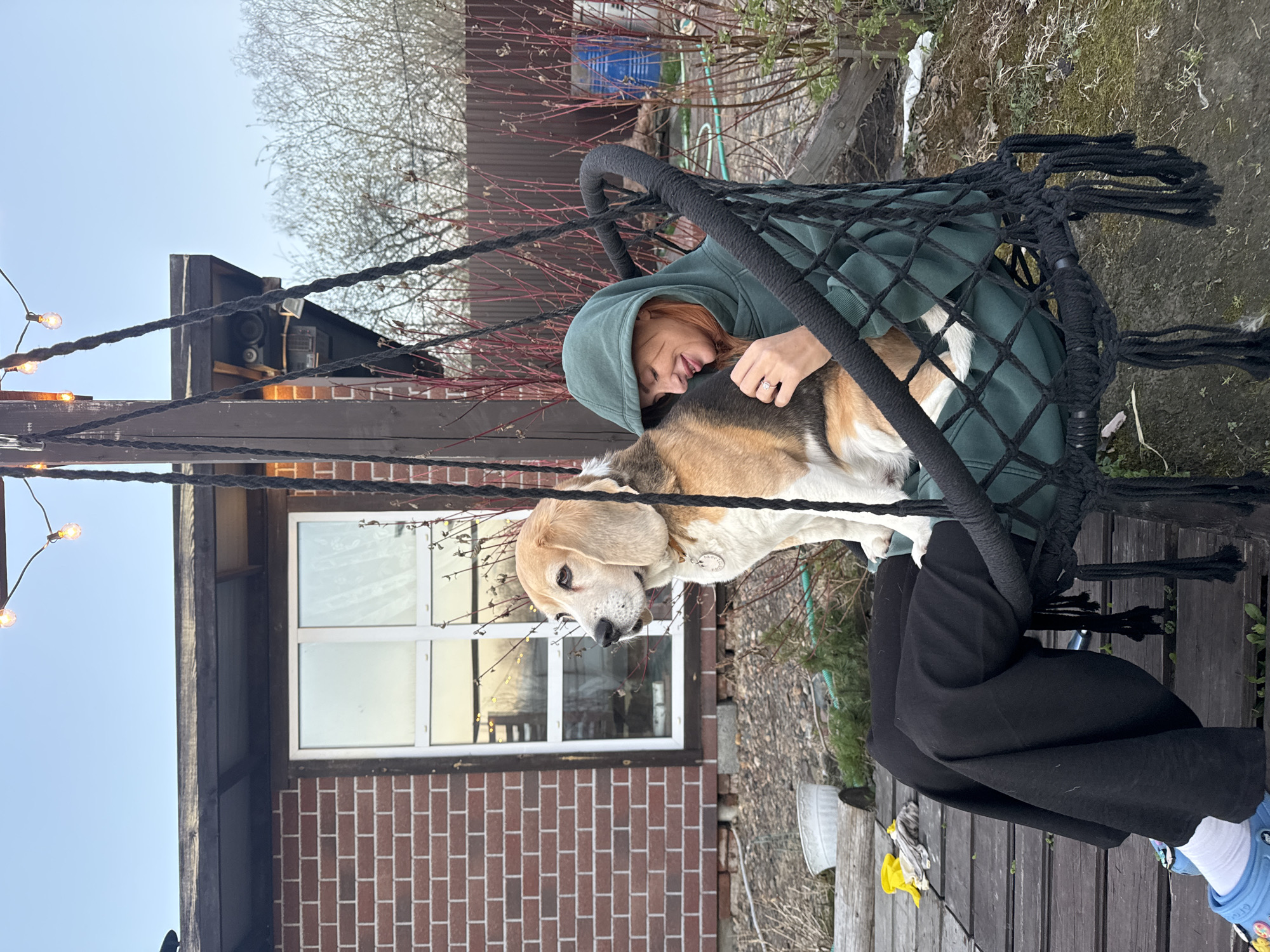 A woman in a green hoodie sits on a black rope swing outdoors, holding a beagle dog. The scene takes place on a wooden deck, with a brick building and leafless bushes in the background. Strings of lights are strung up above. The sky is overcast. The location is Emel'yanovo, Russia.