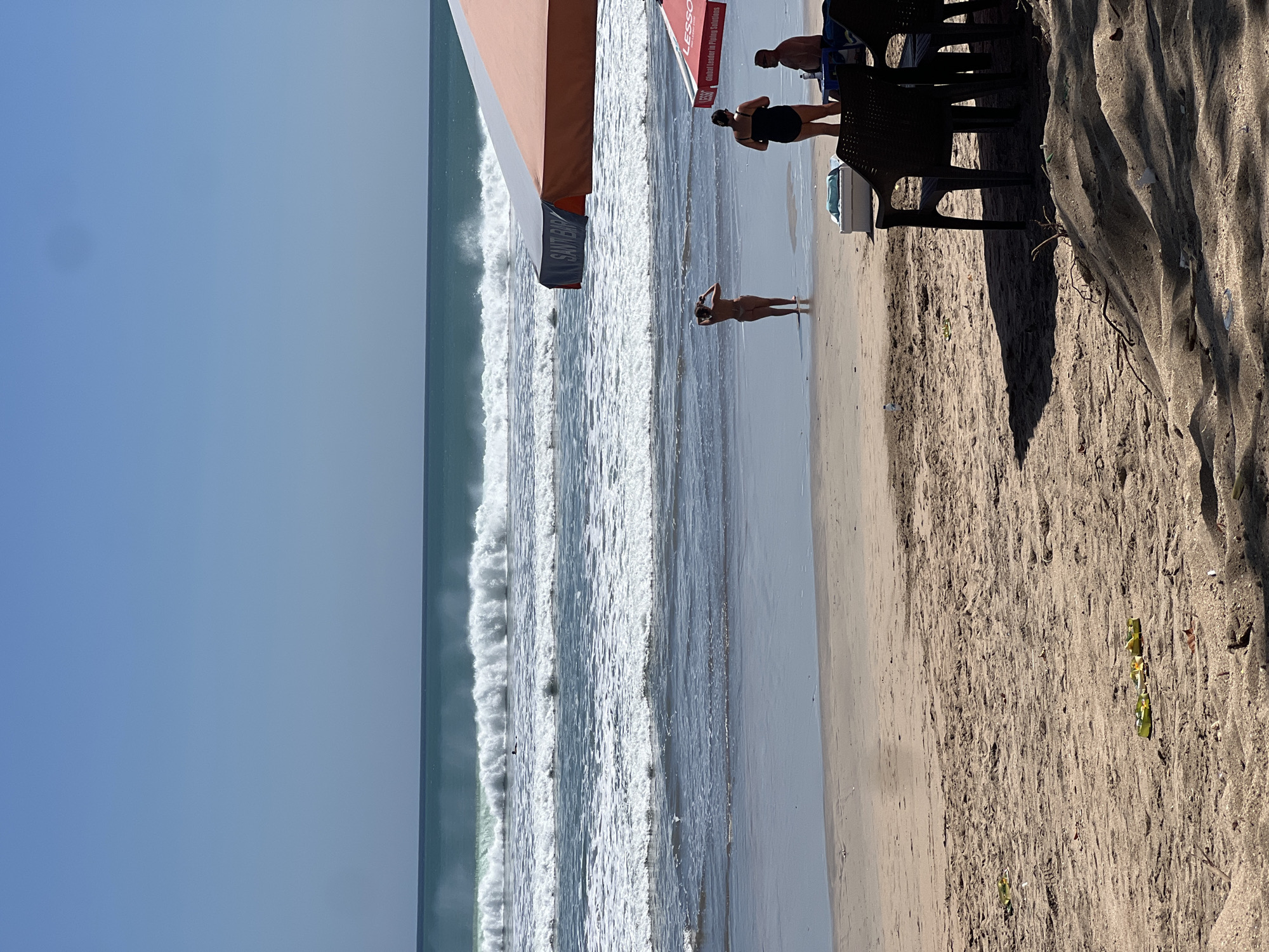 In Kuta, Indonesia, a beach scene unfolds under a clear blue sky. The ocean is rough with large waves crashing, and a lone surfer is visible in the distance. Several people are present on the sandy beach. In the mid-ground, a woman in a swimsuit stands facing the ocean, taking a photo with her phone. Further to the right, another woman in a black swimsuit stands near a man sitting on a chair. Two beach umbrellas provide shade: one is large and orange and white, with text that reads "SANT BAR," and the other is smaller and red, with "LESSO" and "global Leader in Flying Solutions" printed on it. In the foreground, several dark chairs are set up on the sand, some casting shadows. A white cooler is positioned near the chairs, and scattered across the sand are some yellow flip-flops and other debris.