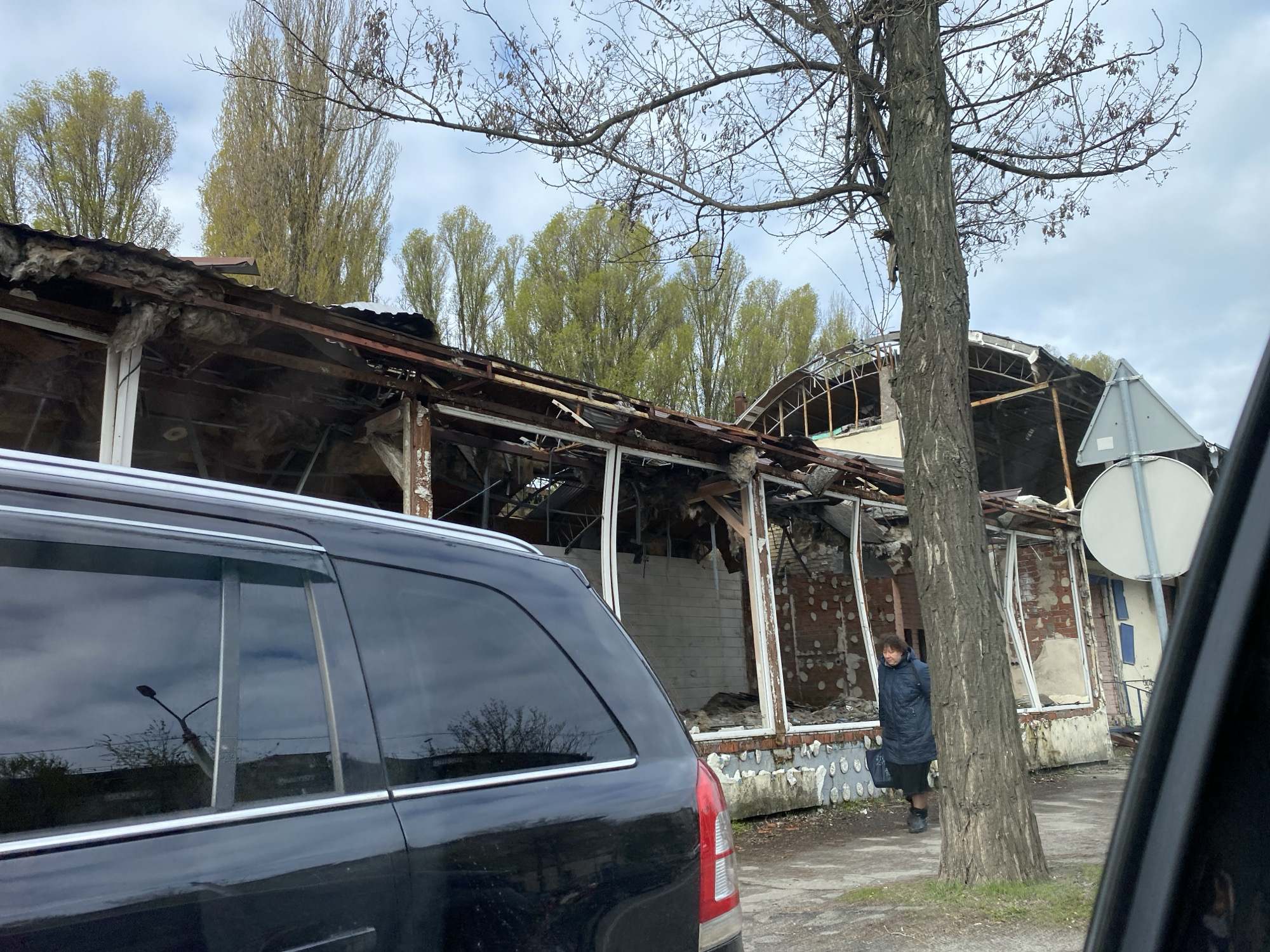 The image depicts the ruins of a building, possibly a commercial structure, with extensive damage to the roof, walls, and windows. The building's frame is still visible, indicating it was likely made of metal and brick. Debris and insulation materials are scattered around the site. A woman in a dark coat stands near the building, beside a tree. A black vehicle is partially visible on the left side of the frame, and part of a vehicle is visible on the right side of the frame, suggesting the photo was taken from inside of a car. A road sign is visible in front of the building. The background features trees with budding leaves, suggesting it is springtime. The location appears to be in the city of Unknown, Unknown.