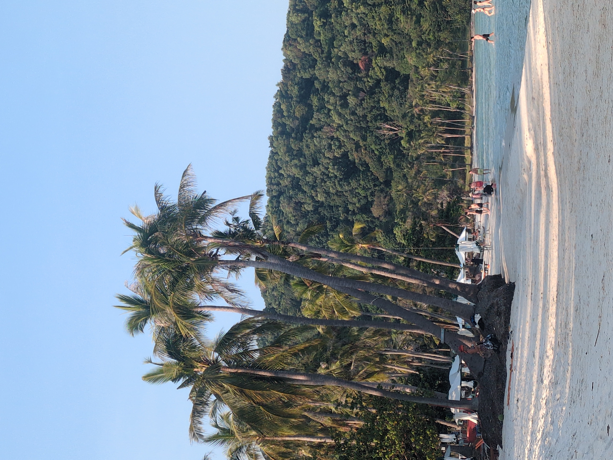 The image shows a beach scene in Phú Quốc, Vietnam. The beach has white sand, clear water, and a lush green hillside as a backdrop. There are several palm trees along the beach edge. People are present in the water and on the beach. Some appear to be swimming or wading, while others are near sunbeds or umbrellas. There are boats anchored in the distance.