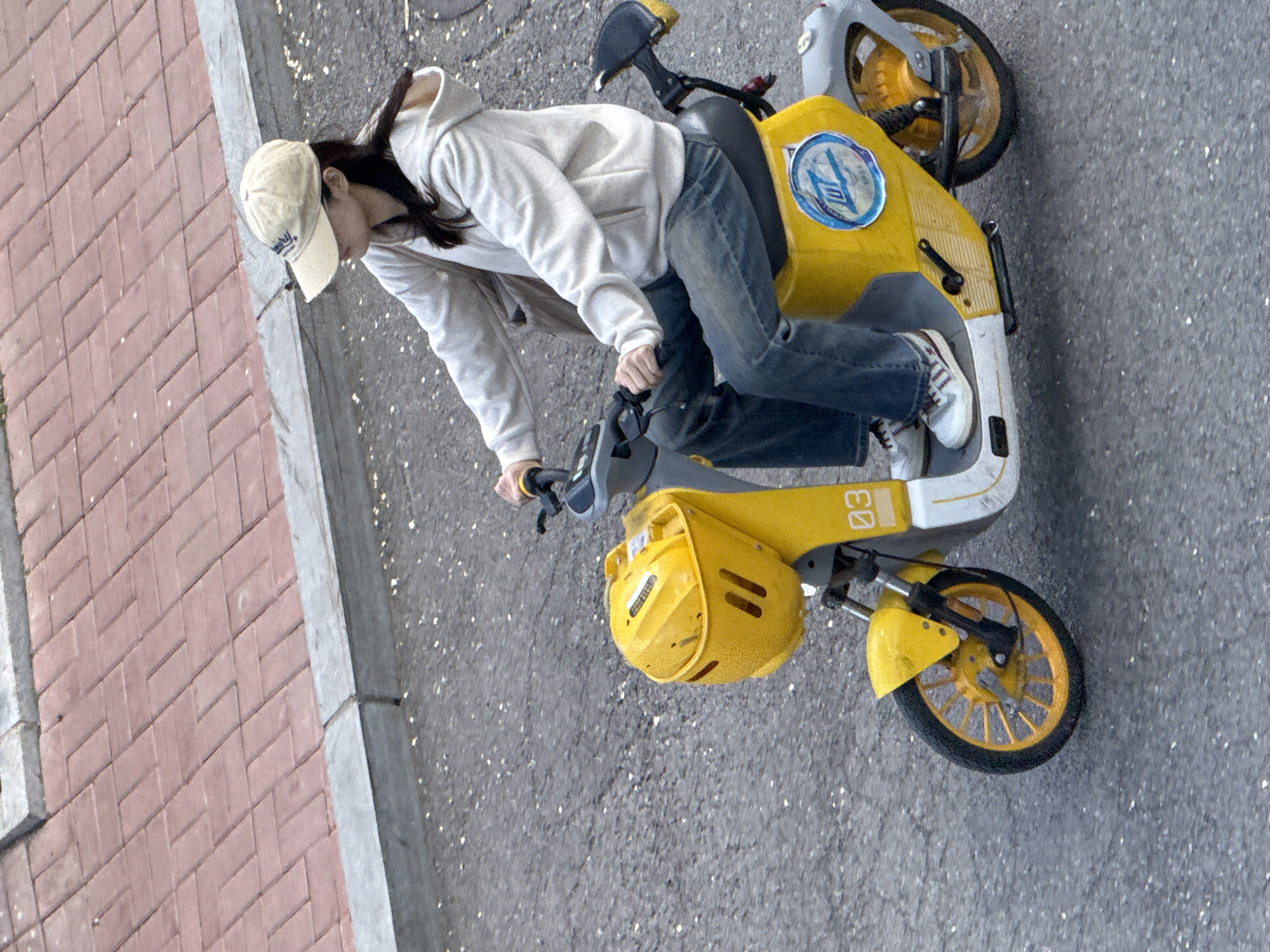 A person wearing a tan baseball cap, a light-colored hoodie, and blue jeans rides a yellow and white electric scooter on a paved surface. The scooter has yellow wheels and a yellow basket on the front. The scooter has a circular emblem. The pavement is grey with some white specks. To the left of the pavement is a brick walkway with grey borders. The location is likely Zhongtun, China.