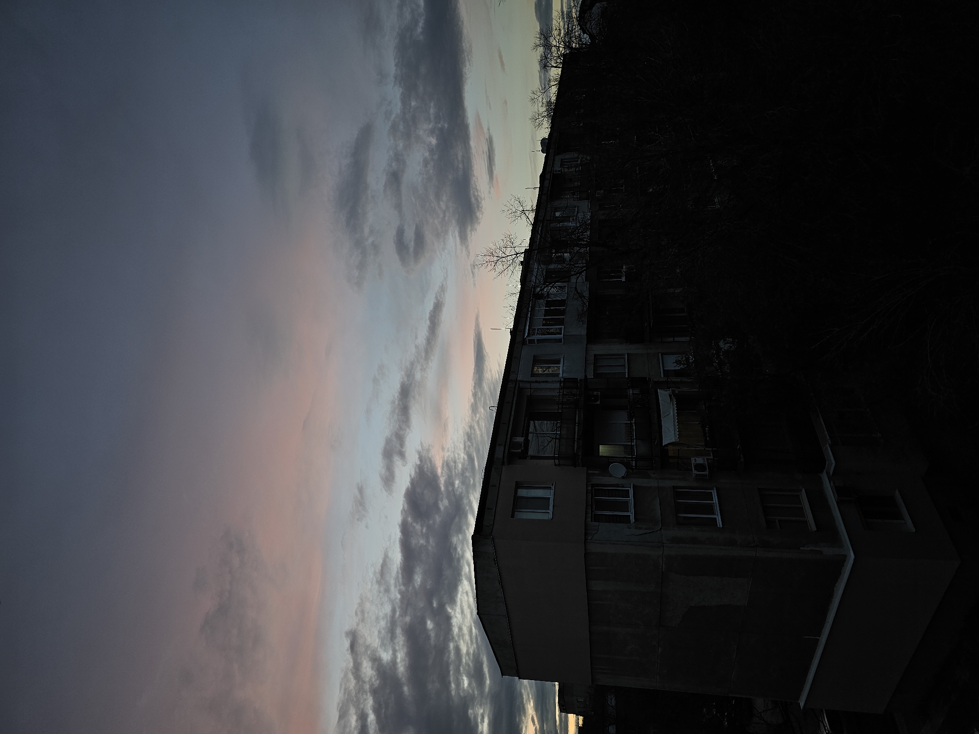 The image shows a multi-story residential building in Plovdiv, Bulgaria at dusk. The building is faded and appears somewhat dilapidated. It has a mix of visible windows, some with air conditioning units. A satellite dish is mounted on one of the walls. Trees, bare of leaves, are adjacent to the building. The sky is filled with clouds tinged with pink and blue hues from the setting sun. The right side of the image is very dark.