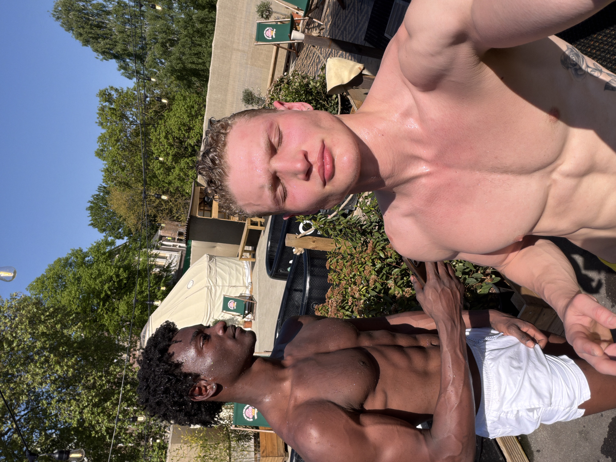 This outdoor shot features two muscular men on a bright, sunny day. The man in the foreground, with fair skin and light curly hair, looks directly at the camera with his eyes slightly closed, suggesting relaxation or squinting against the sun. His upper body is shirtless, showcasing a well-defined physique, and his skin appears wet or glistening with sweat. Behind him, a second man with dark skin and dark curly hair is also shirtless, displaying a similarly athletic build. He wears white shorts and gazes upwards and away from the camera, his skin also wet.
The scene takes place in what appears to be a recreational or wellness area, possibly a pool deck or outdoor event space. The background features a clear blue sky, green trees, and hanging string lights. Light-colored tent-like structures or awnings are visible further back, along with several green folding chairs. These chairs bear a circular logo, which is difficult to read precisely but appears to be tennis-related, possibly associated with Roland Garros. A black tub or plunge pool is partially visible behind the dark-skinned man, suggesting it might be an ice bath or similar facility. The strong, direct sunlight indicates it is midday or early afternoon, casting distinct shadows. The overall impression is one of post-activity relaxation or a casual outdoor gathering.