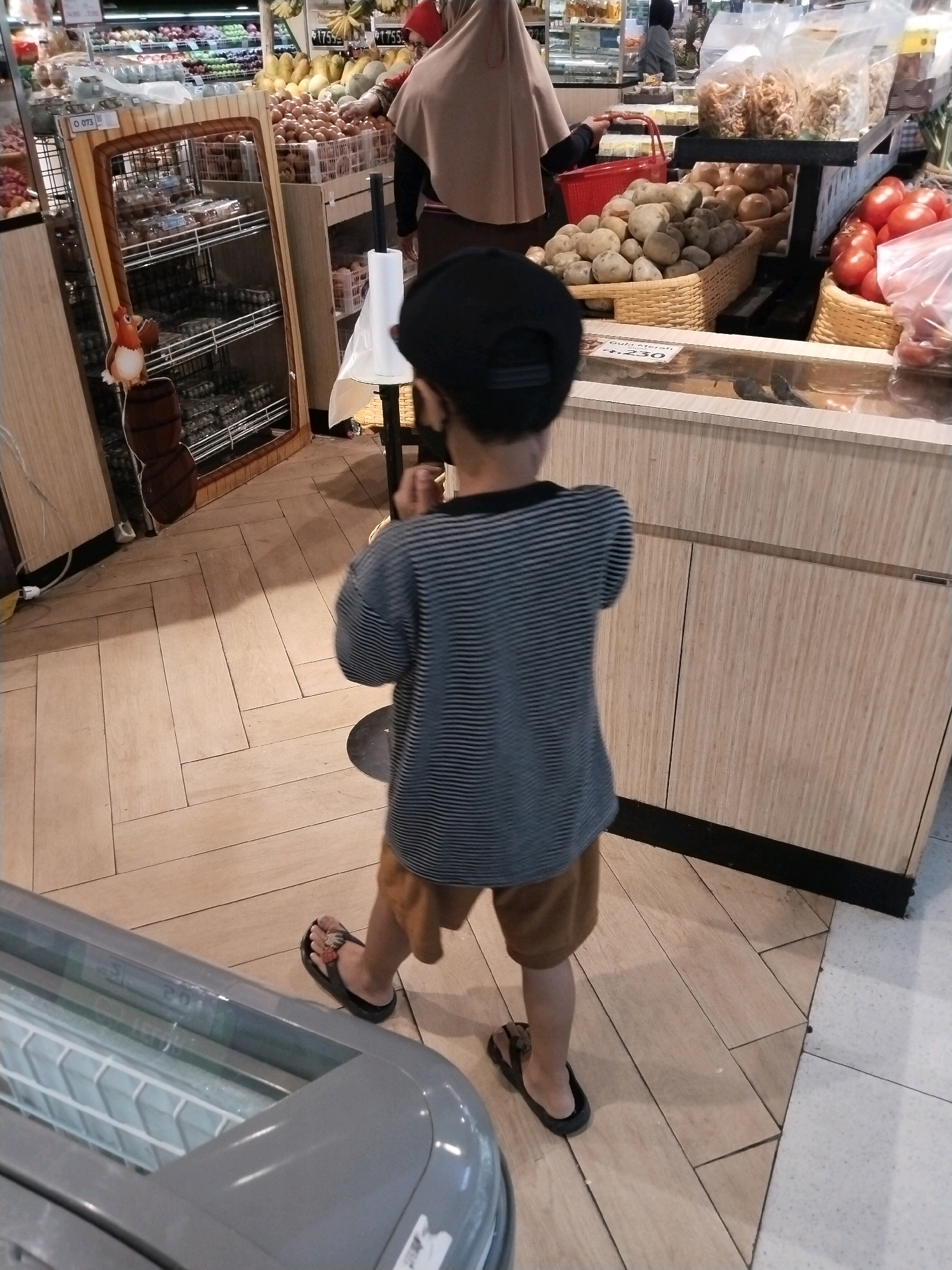 A young child wearing a black cap, a striped long-sleeved shirt, and brown shorts is standing in an aisle of a supermarket in East Jakarta, Indonesia. The child is facing away from the camera and is partially obscured by a refrigerator unit in the foreground. In the background, a woman wearing a hijab is visible, along with displays of produce and packaged goods. The floor is made of light-colored wooden planks laid in a herringbone pattern.
