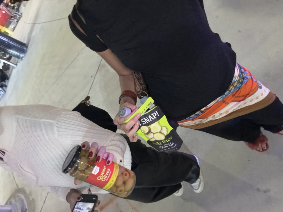 Two people stand next to each other. One person holds a glass jar of “Queen Olives”, and the other holds a bag of “That’s SNAP! Zilly Bites Dill Pickle Sliced Pickled Cucumbers”. One is wearing a white sweater and black pants, and the other is wearing a black shirt and black pants with a colorful scarf around the waist.