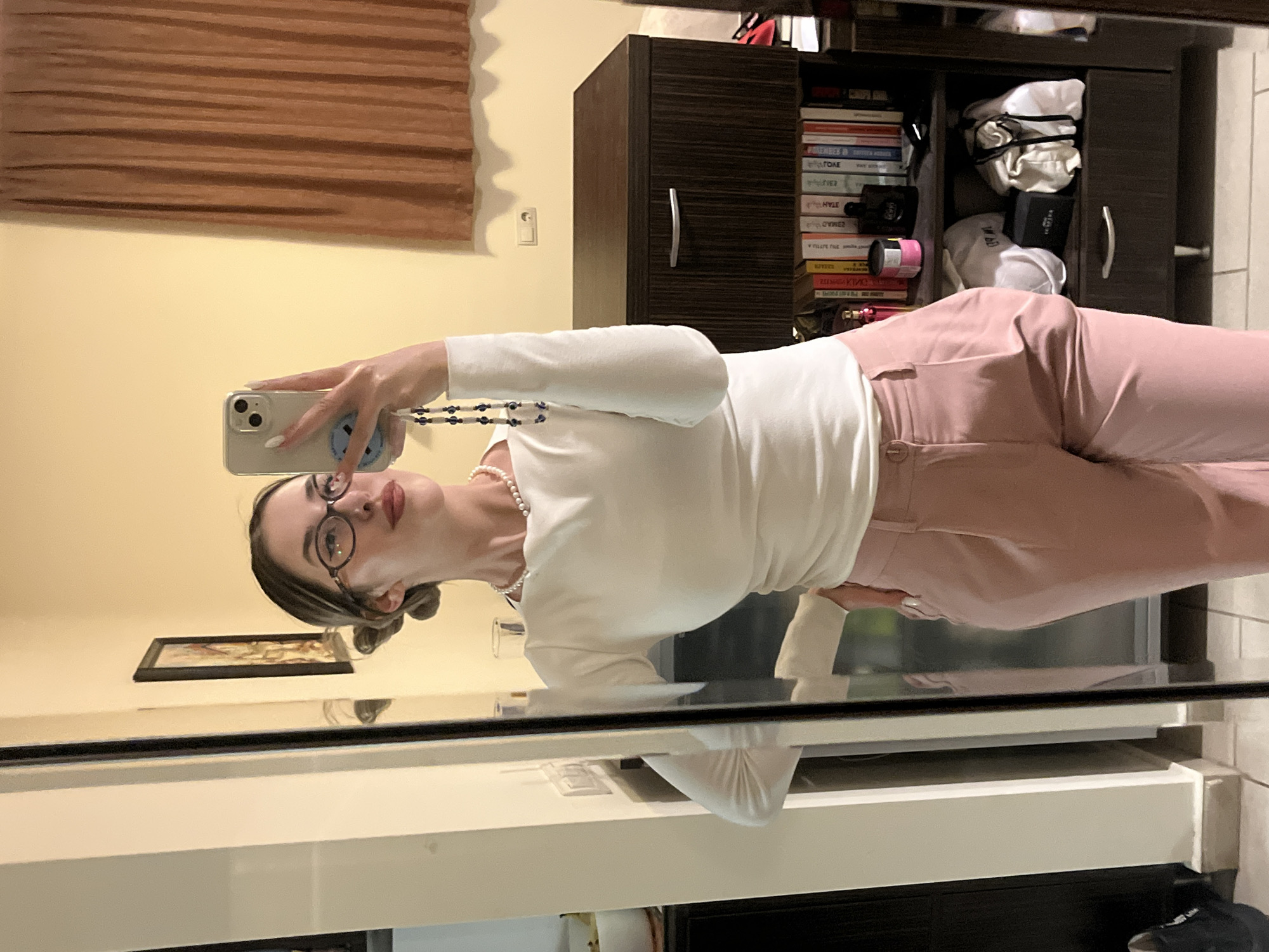 An indoor mirror selfie shows a young woman in Sofia, Bulgaria, posing in front of a mirror. She is holding an iPhone with a light-colored case and a blue and white patterned strap to capture her reflection. She wears a long-sleeved white top, light pink trousers, round-framed glasses, and a pearl necklace. Her hair is styled in low buns, and her fingernails are painted white.
The background features a warm-toned (yellowish-beige) wall with a light switch or outlet. To the left, a horizontal wooden blind or curtain is partially visible. To the right, a dark brown wooden cabinet or bookshelf is filled with books, some showing partially legible or reflected text on their spines (e.g., "TOAE", "HEO", "ZUMA"). White and black objects, possibly bags or boxes (one showing "The BIG" and another "NIXON"), are also on the shelves. A framed picture hangs on the wall below the woman's head in the reflection. Below the main mirror frame, a white surface, possibly a counter or table, is visible, reflecting parts of her outfit. The scene suggests a home setting, possibly in the late afternoon or evening, given the indoor lighting. The woman maintains a calm, posed expression for the selfie.