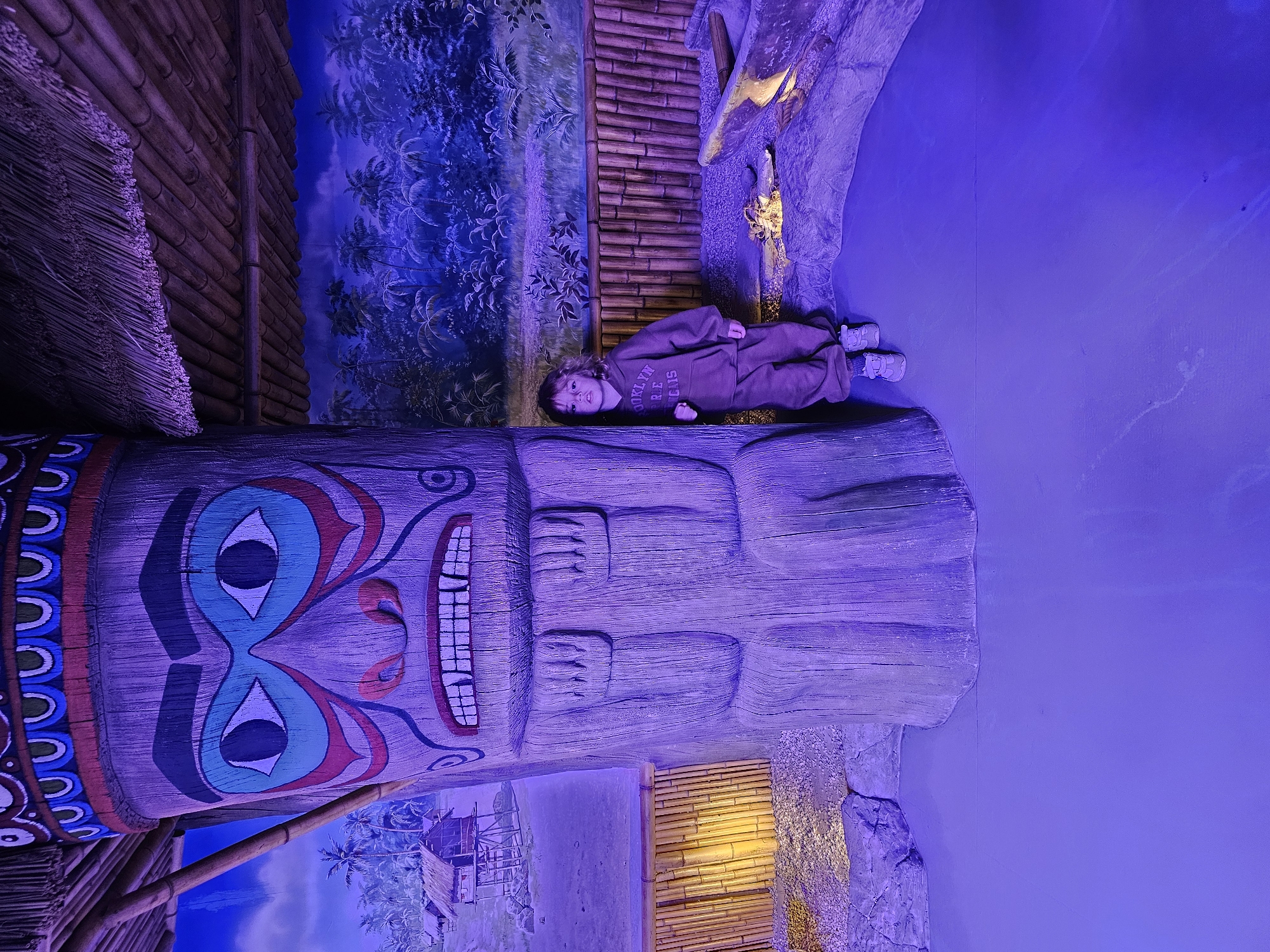 A child in a brown jumpsuit lies on a large wooden totem pole with a painted face in what appears to be an indoor, themed environment. The background includes a painted mural of a tropical landscape with bamboo structures. This is in Khimki, Russia.