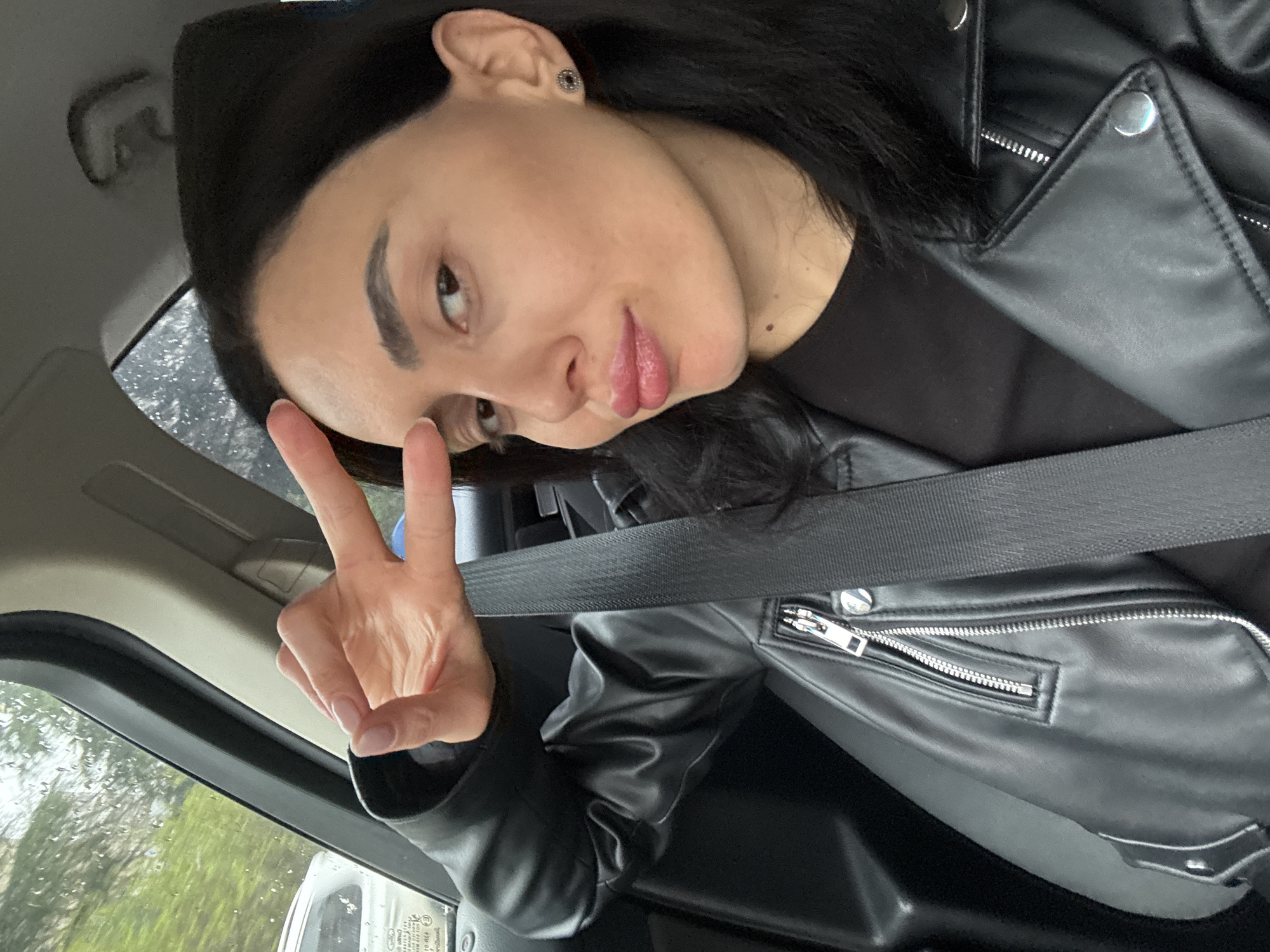 A young woman is in a car, wearing a black leather jacket and a seatbelt. She is holding up a peace sign. It's raining outside, and the view from the car window shows greenery, suggesting an area with trees or parks. The scene is most likely in a car in Tbilisi, Georgia.