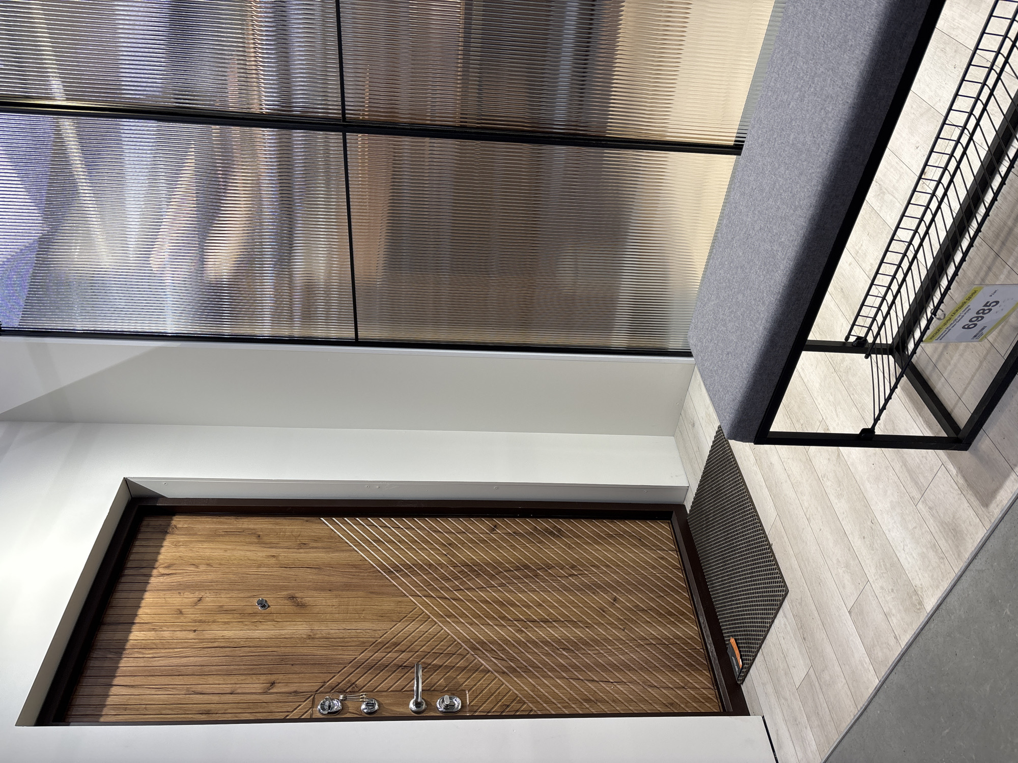 A high-angle shot depicts the interior of what appears to be a building in Perm, Russia, exhibiting a blend of modern design elements. The focal point is a wooden door, inset within a white wall, detailed with diagonal linear patterns and a metallic handle and locking system. Above the door, a window with a black grid pattern and textured glass offers a view to the outside.
Adjacent to the window is a gray bench with a black metal frame, angled towards the right side of the frame. A small, rectangular gray mat is positioned in front of the door. The floor is covered with light-colored, wood-patterned tiles, leading to a gray surface on the right.
The overall environment suggests an entryway or waiting area, characterized by a mix of textures and geometric lines, indicative of contemporary interior design.
