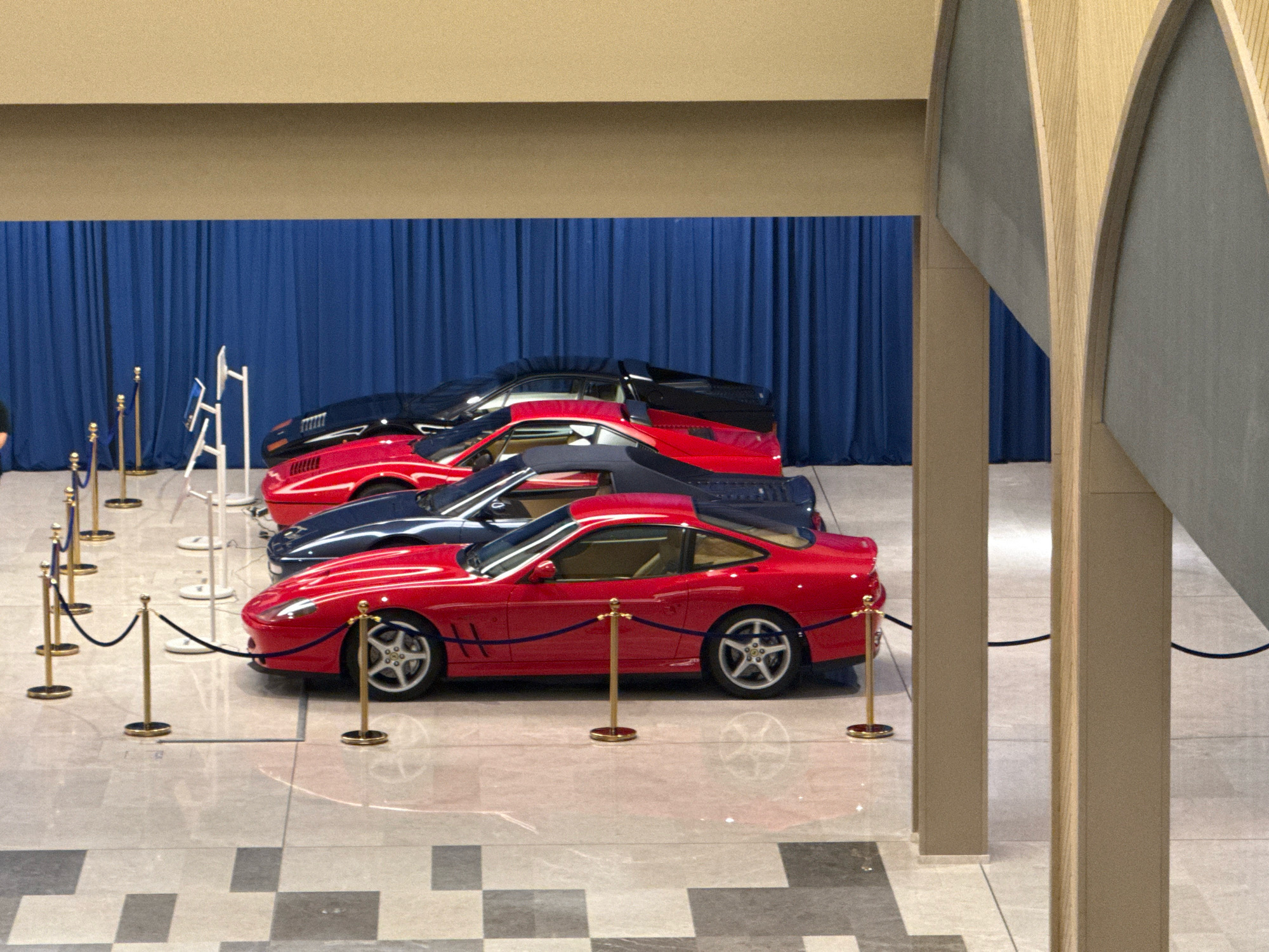 The image shows a row of luxury sports cars, primarily red and dark blue, displayed indoors behind velvet ropes and stanchions. The cars are parked on a polished floor with a checkered pattern. In the background, a dark blue curtain runs along the wall. The scene appears to be a car exhibition or showroom. The architectural elements, with their curved wooden beams, suggest a modern building. The lighting is bright and even, illuminating the vehicles and the surrounding space. The location is identified as the Tashkent International Airport in Tashkent, Uzbekistan.
