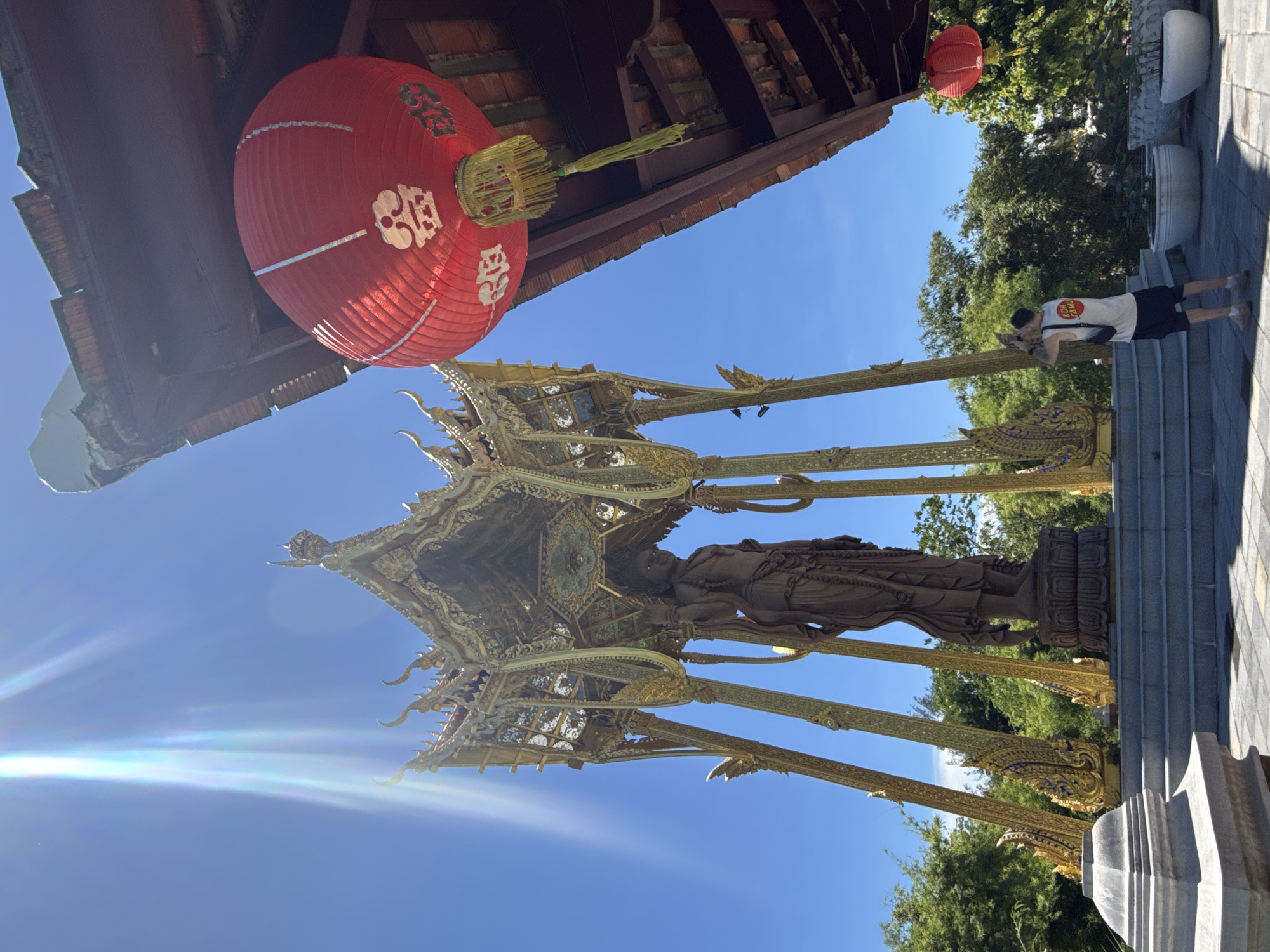 A person is photographing a golden statue of Buddha. The statue is situated in a golden, ornate pavilion-like structure. A red Chinese lantern is visible in the upper left of the frame. The environment is bright and sunny, suggesting daytime. The statue and lantern are in Bang Pu Subdistrict Municipality, Thailand.