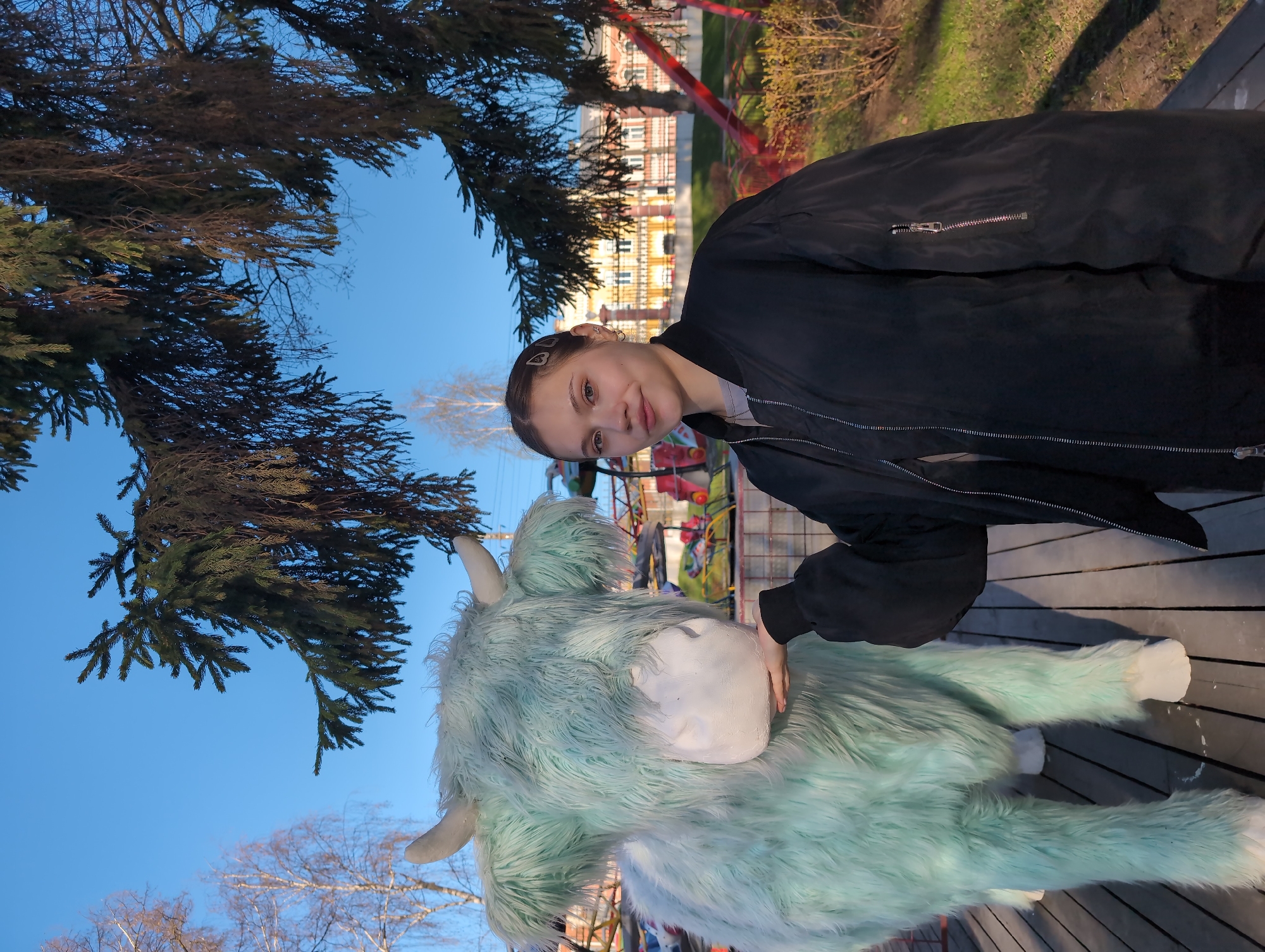 A young woman in a black jacket poses with a light blue, furry bovine sculpture on a wooden platform. In the background, urban elements are visible: a playground with red support structures, a building with orange and white facade details, and deciduous and evergreen trees under a clear blue sky. The location is Izhevsk, Russia.