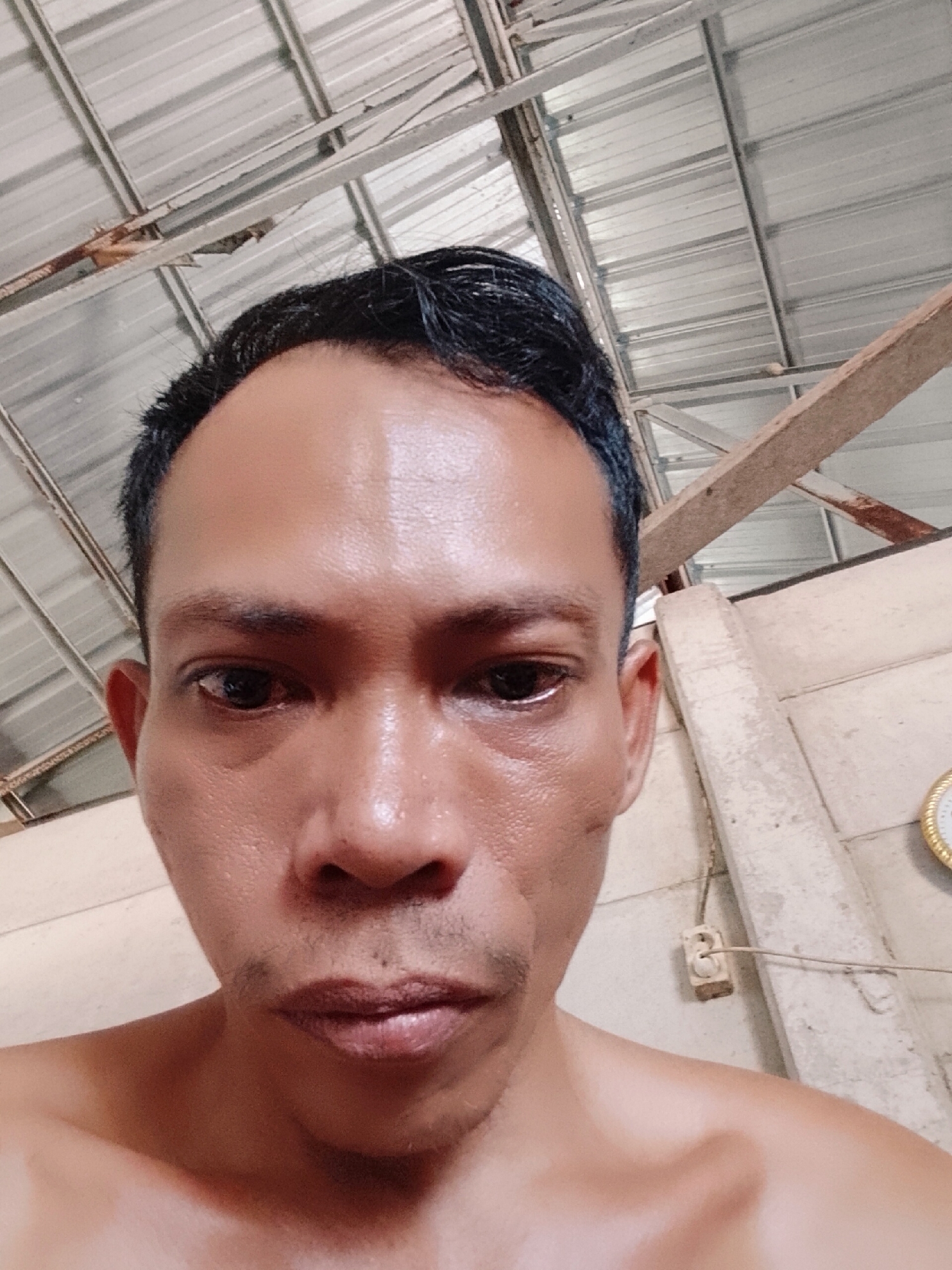 The image shows a man with short black hair and a bare upper torso. His skin is moist and he has dark eyes. He is looking directly at the camera. The background includes a corrugated metal ceiling, wooden beams, and a wall with an electrical outlet and a clock. The location is Bukit Ketok, Indonesia.