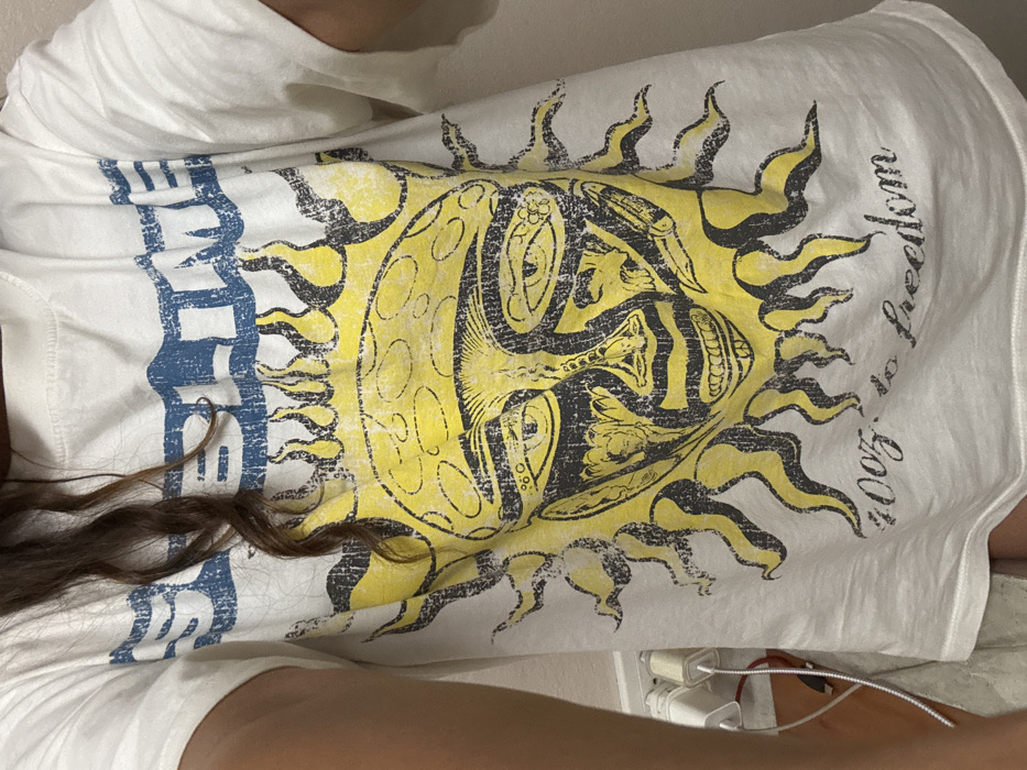 A white T-shirt with a large yellow and black sun graphic dominates the image. Inside the sun's center is an intricate design with abstract figures. "2007 to freedom" is printed in a circular arc near the sun's edge. A horizontal blue and white design, possibly letters, sits left of the sun. A small portion of brown wavy hair falls across the shirt. Electrical outlets and charger plugs are visible on the bottom right.