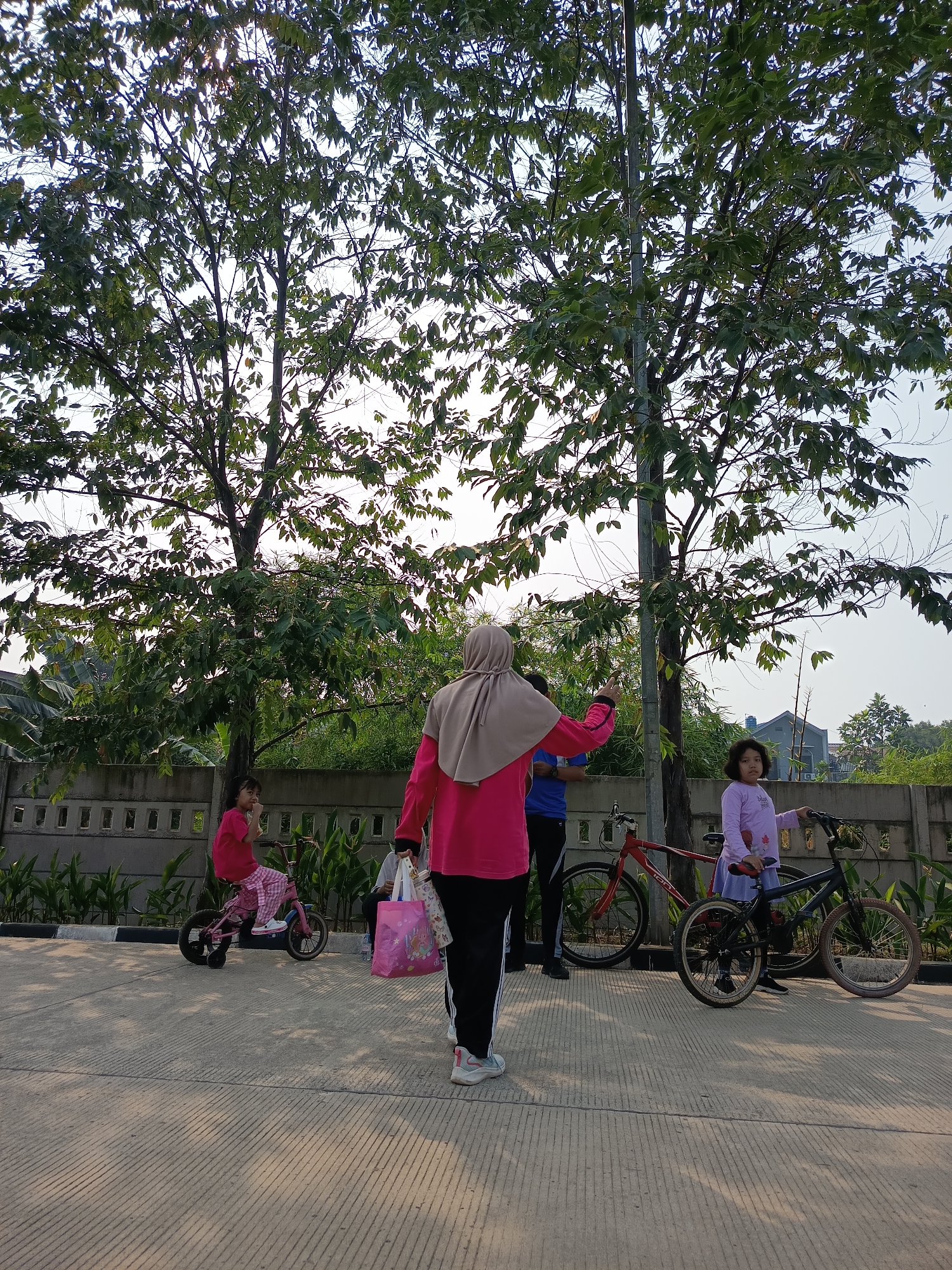 In Bekasi, Indonesia, on a paved pathway lined with concrete planters and lush green foliage, two trees with full canopies dominate the background under a clear sky. In the foreground, a woman in a pink top and hijab, holding a pink shopping bag, walks away from the camera. To her left, a young girl in a pink top and checkered pants rides a pink bicycle with training wheels. Further to the right, another young girl in a light purple long-sleeved shirt and skirt stands next to a black bicycle. A man in a blue shirt and black pants is partially visible between the two women. In the background, two bicycles, one red and one black, are parked. The scene appears to be a park or recreational area, given the presence of bicycles and people engaged in outdoor activities.