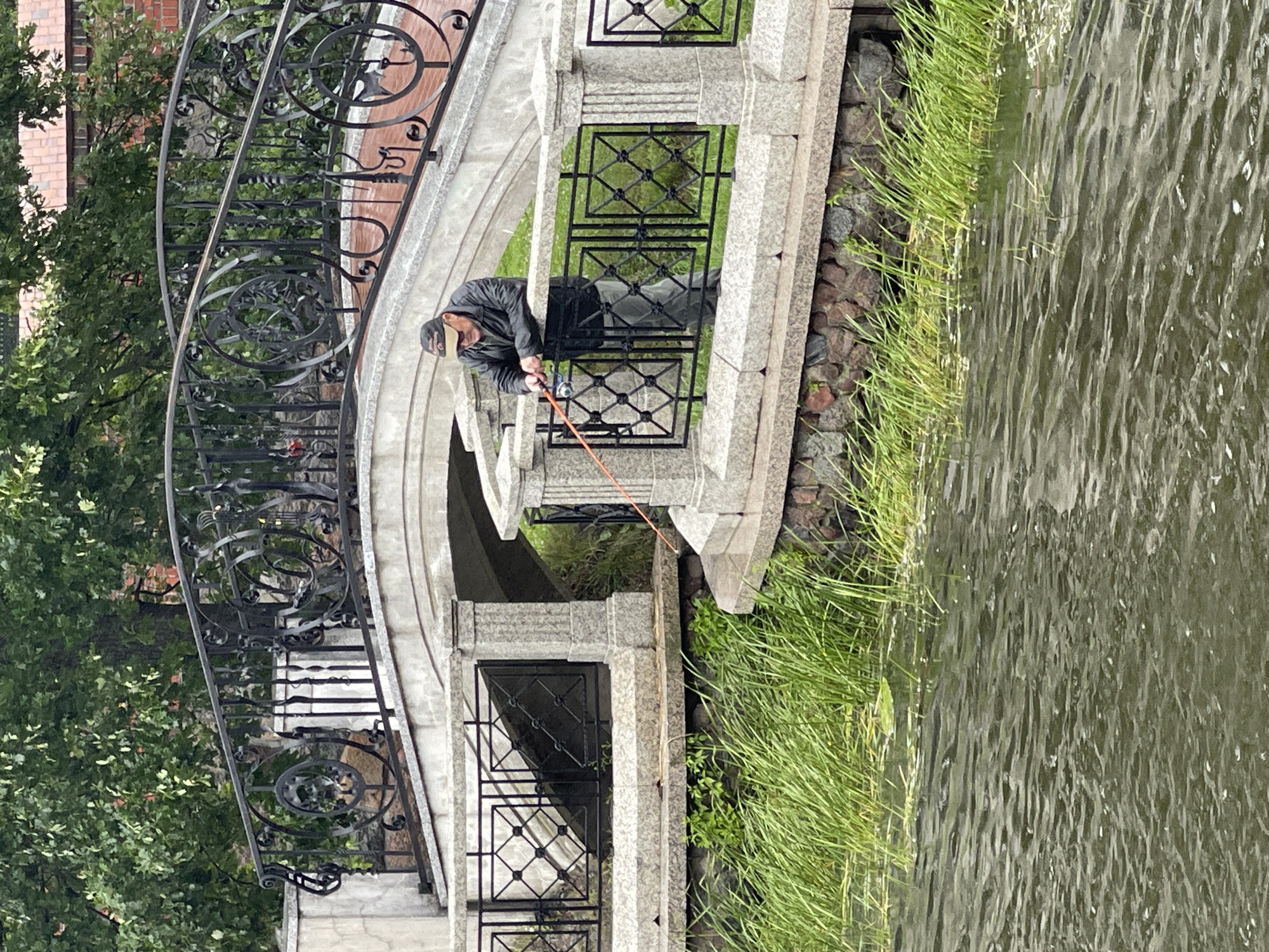An overhead shot captures a man fishing from the edge of a stone embankment. He stands with his back to the viewer, leaning on a decorative metal railing that borders a walkway. The man wears a dark jacket, grey pants, and a baseball cap. He holds a fishing rod, its tip extending into the murky water below. The embankment is lined with lush green grass and a stone retaining wall. In the background, a picturesque arched bridge with ornate black metal railings spans the waterway. Trees with dense green foliage and a glimpse of a red-tiled roof suggest an urban park or historic setting. The water is a dull green, rippled by a gentle current. The scene appears to be along the Pregolya River in Kaliningrad, Russia, possibly near the historic Fishing Village.