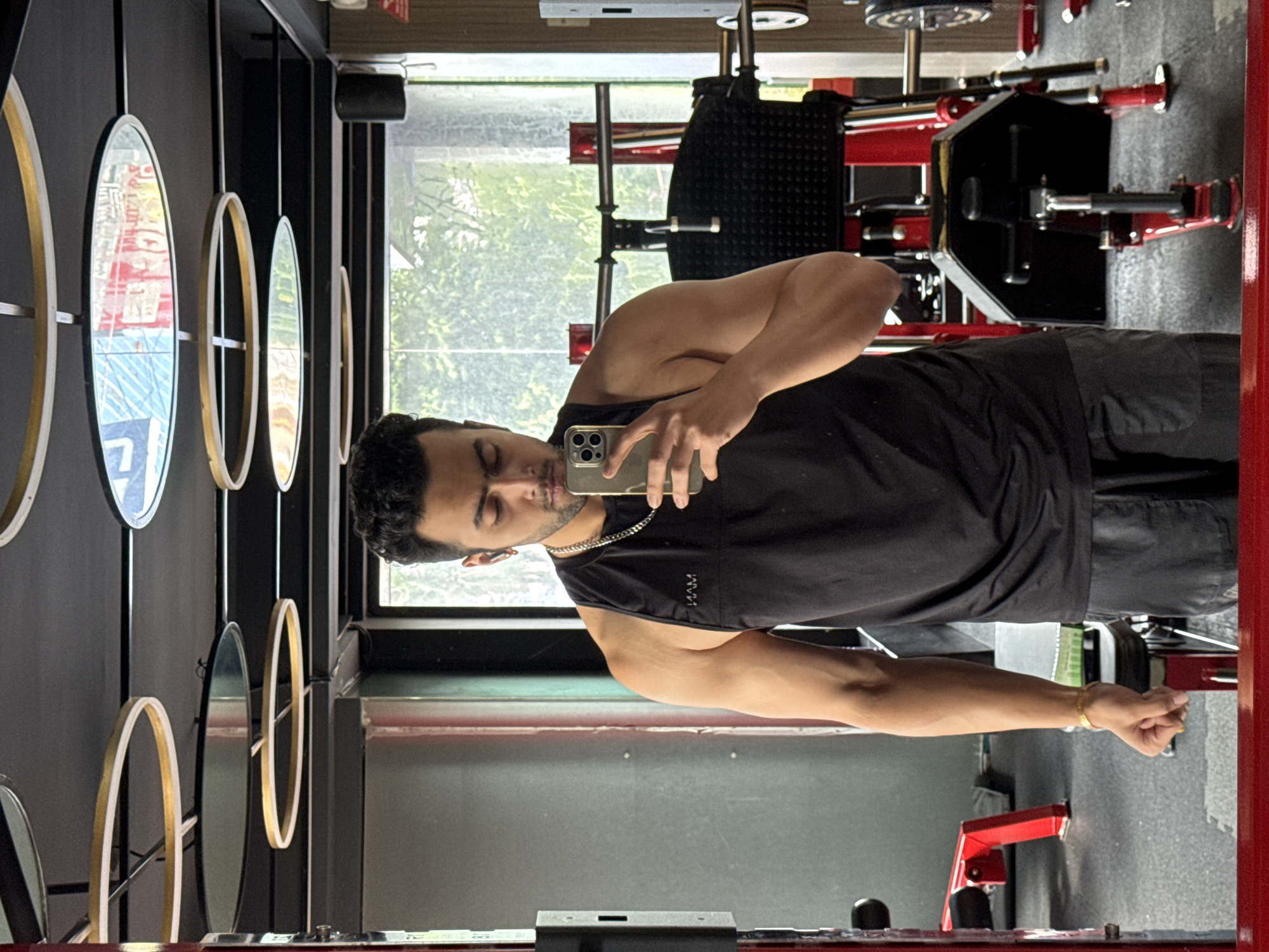 In Kolkata, India, a man with dark, curly hair and a beard takes a mirror selfie in a gym. He is wearing a black sleeveless t-shirt and dark grey shorts, with a silver chain around his neck and earbuds in his ears. His left arm is flexed, and he holds a smartphone in his right hand. The gym features a grey floor, a large mirror reflecting the man and the gym equipment, including red and black weight machines and a glimpse of trees and sky through a window. The ceiling has a modern design with circular lights.