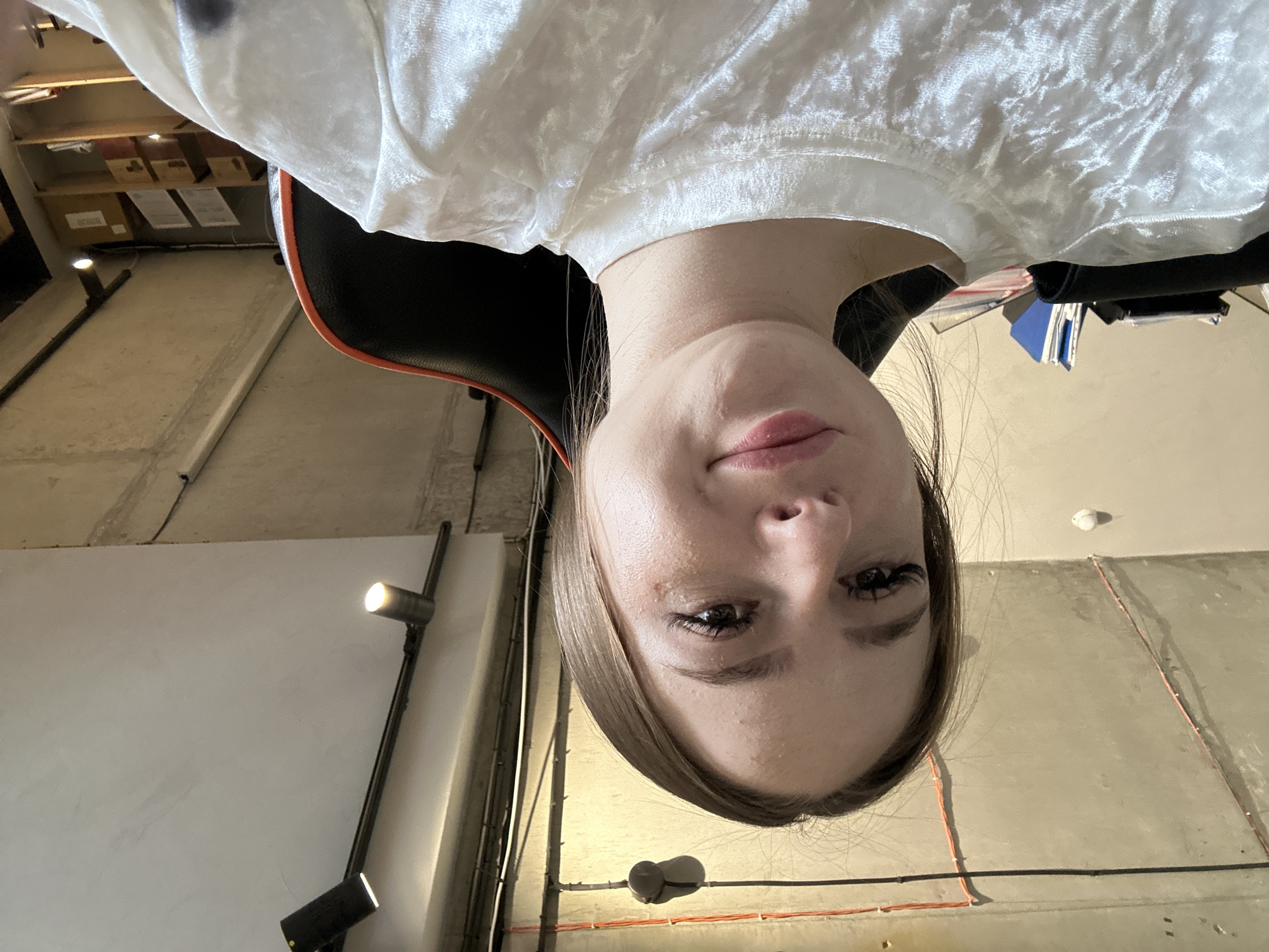 A young Caucasian woman with brown hair pulled back is captured in a selfie from the chest up, indoors. She is wearing a white, textured shirt and is seated in a black and orange gaming chair. The background reveals a raw concrete ceiling with visible electrical conduits and lighting fixtures. To her left, a stack of folders and papers is on a surface. The scene appears to be in an office or studio environment with unfinished architectural elements.