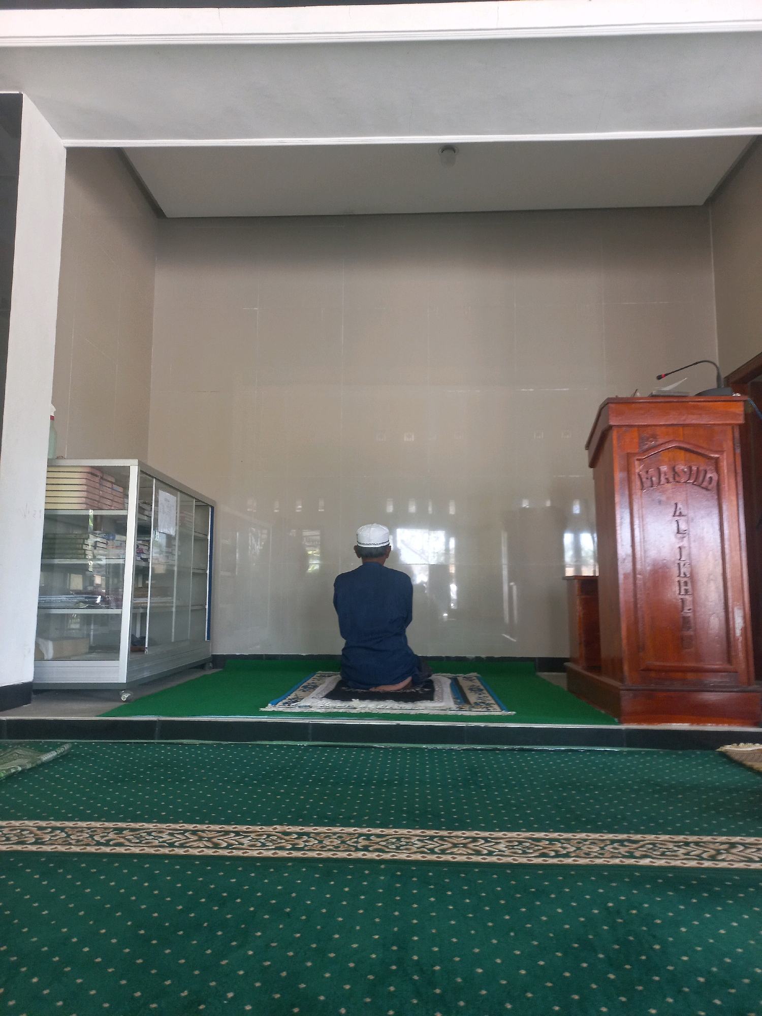 The image shows a man wearing a white cap and a dark blue long-sleeved shirt, kneeling on a prayer mat in what appears to be a mosque. He is facing away from the camera, engaged in prayer. The prayer mat is placed on a green carpeted floor. In the background, there is a glass display cabinet filled with books, possibly religious texts. To the right, a wooden podium stands, also with some inscription. The walls of the room are tiled in a light, neutral color, and there are windows or reflective surfaces that show the exterior. The lighting suggests it is daytime, possibly indoors with artificial lighting. The overall scene conveys a sense of tranquility and devotion. The location context provided is Nguntoronadi, Indonesia. The visible text on the podium reads "MASJID AL-IKHLAS".