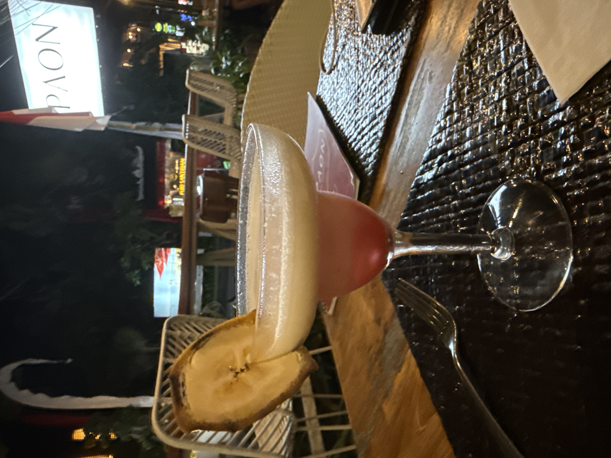 A close-up shot focuses on a cocktail in a margarita glass, garnished with a dehydrated banana slice. The drink has two distinct layers, pink on the bottom and a creamy white on top, with a foamy head. The glass sits on a wooden table with a textured placemat. A fork is visible to the left of the placemat. In the background, out of focus, are wicker chairs, a sign that reads "PAON", and a flag. The scene appears to be an outdoor dining area at night, likely in Denpasar, Indonesia, suggested by the signage and tropical ambiance.