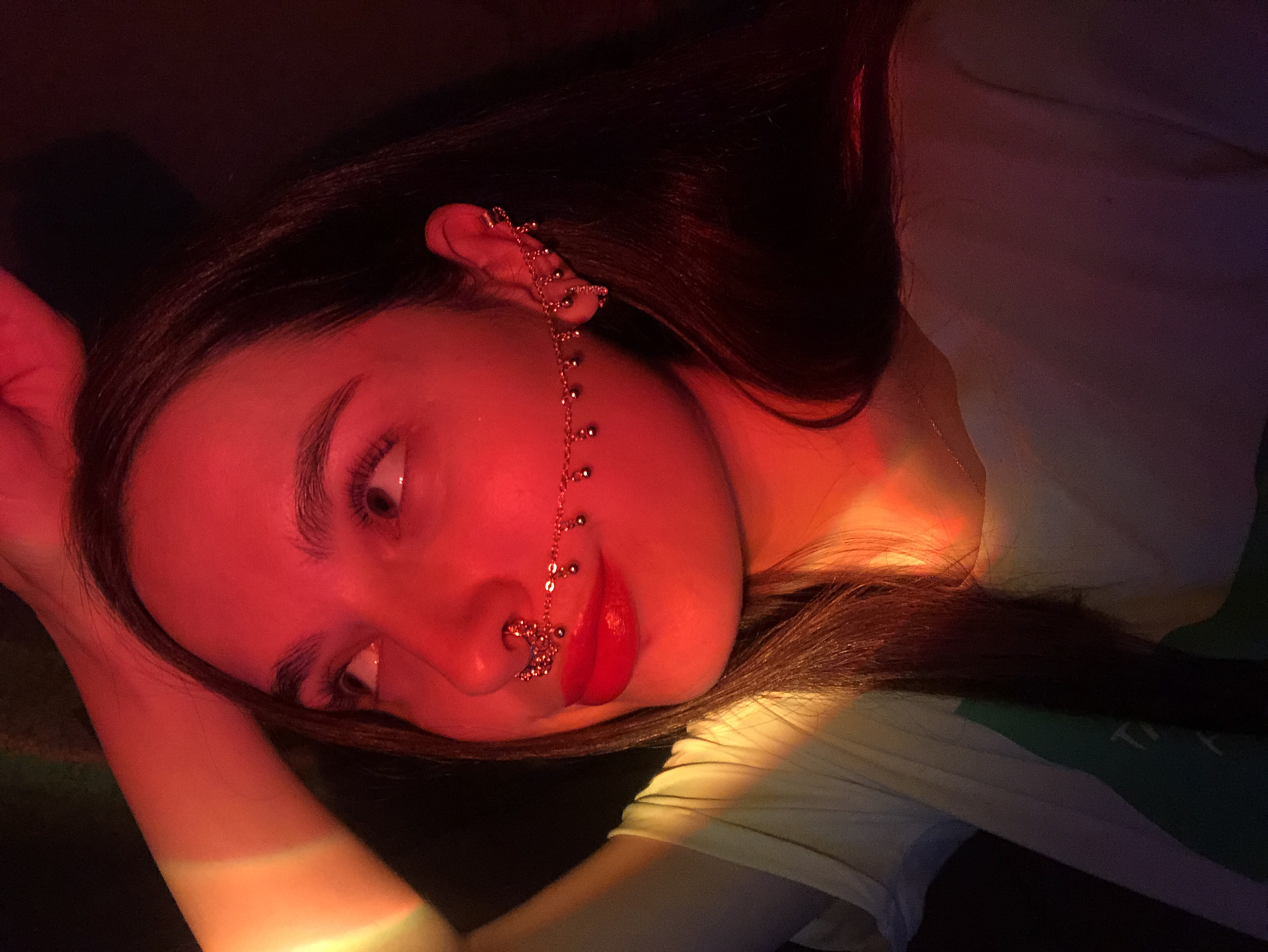 A woman with long, dark hair is lying down, facing the camera. She is wearing a nose ring and a chain connecting her ear to her nose ring. Her lips are painted bright red. The lighting is predominantly red, with some hints of orange and yellow. The setting appears to be indoors. There is no information to locate the scene in the city of Noviy, Russia.