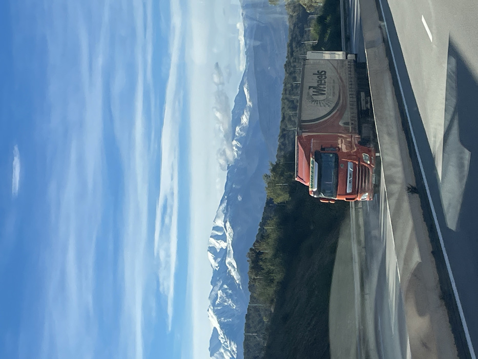 A vibrant blue sky with elongated clouds contrasts sharply with snow-capped mountains. An orange truck with "Wheels" printed on its trailer travels down a highway. The roadway is reflective, showing a hint of scenery and vehicle. Green trees line the edge of the road, positioned between the mountains and the highway.