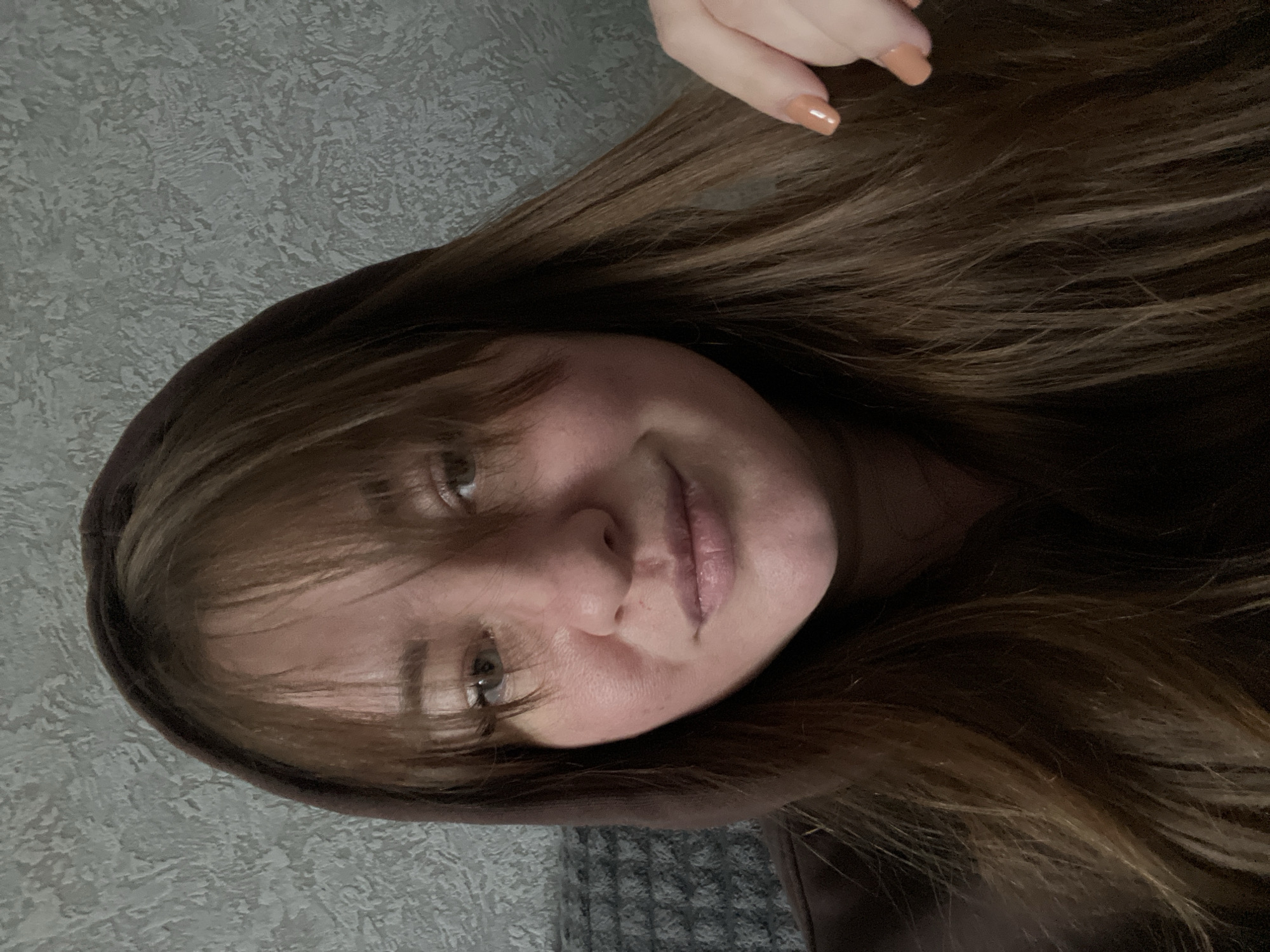A young woman with light skin and long, brown hair with bangs is centered in the image. She is wearing a brown hooded sweatshirt. The background consists of a textured gray wall. The location cannot be determined from the image.