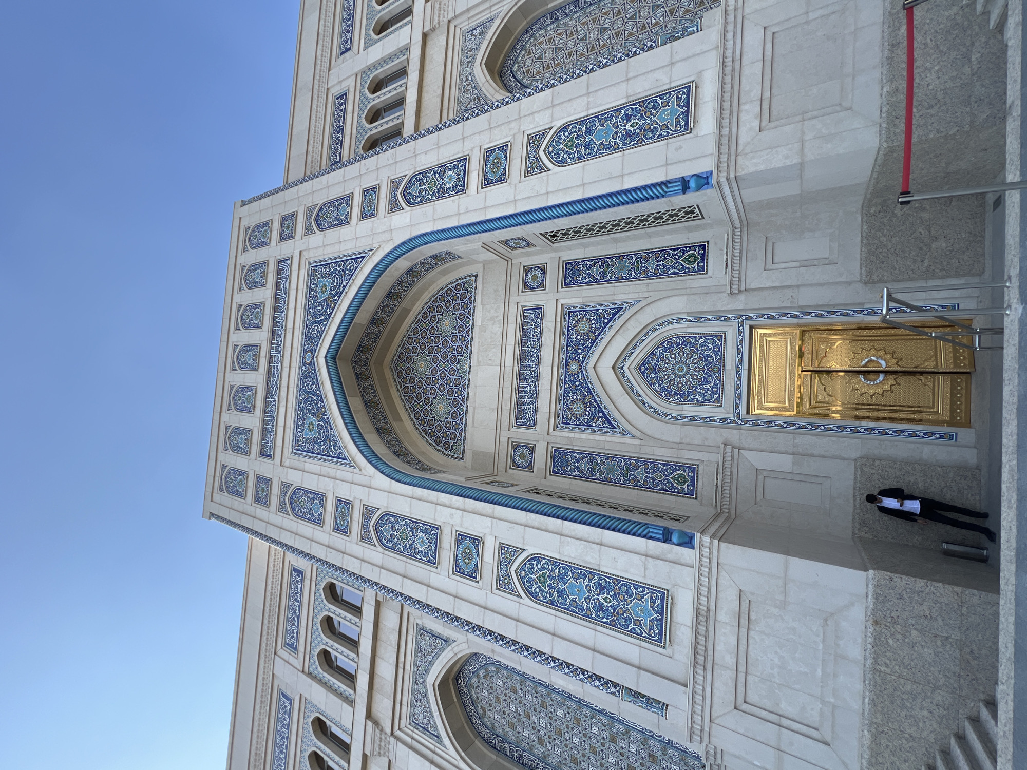 An eye-level, wide-angle shot captures the grand facade of a building in Tashkent, Uzbekistan, likely a religious or administrative structure, under a clear blue sky. The building features intricate blue and white tilework in geometric patterns and Arabic calligraphy adorning its cream-colored stone exterior. A large, ornate golden doorway is centrally positioned, flanked by tall, narrow arched windows with similar tilework. To the left of the doorway, a lone individual dressed in a dark suit and white shirt leans against the building's base, their posture suggesting a moment of pause. Stairs are visible to the far left, leading up to the entrance. A red velvet rope barrier is present on the right side of the frame. The lighting suggests it is daytime, with the sun casting subtle shadows.