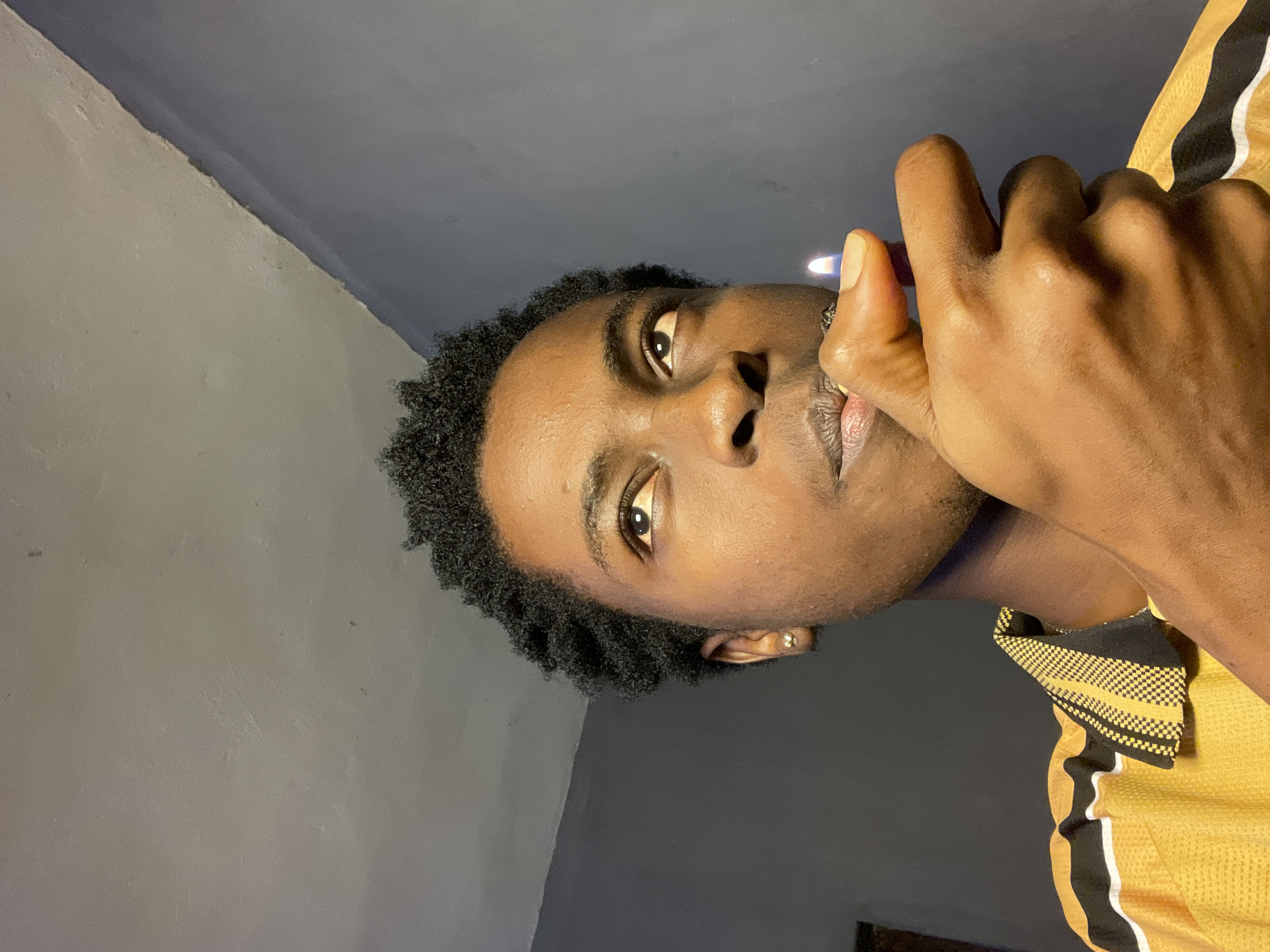 A medium, close-up shot looking up at a young, dark-skinned African male with short, curly black hair holding a lit lighter to his lips. He wears a gold stud earring in his left ear and a gold and black shirt. The background consists of a gray wall meeting a darker gray ceiling. The location is not discernible from the image.