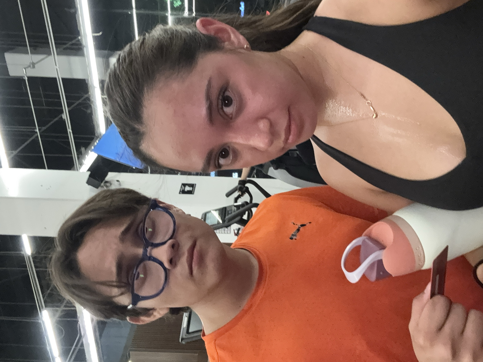 Two people are in a gym. The person on the left is wearing glasses and an orange shirt. The person on the right is wearing a black top. Both appear sweaty. The location is likely a gym but not verifiable as being Puebla, Mexico.