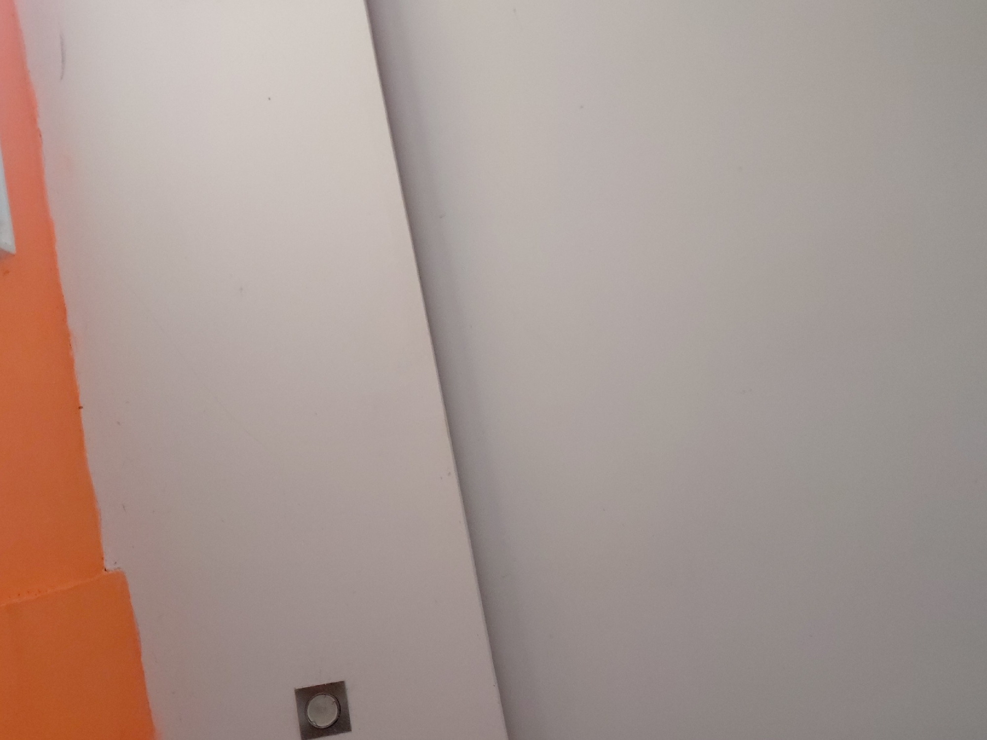 The image displays an interior architectural detail, likely a corner or junction of walls and possibly a ceiling, set in Santa Ana, Venezuela. The dominant feature is a light-colored surface, appearing white or very light grey, which occupies most of the frame. A distinct diagonal line, possibly a corner or a recessed panel, divides this light surface into two planes, with a subtle shadow suggesting depth and separation.
On the far left, a section of a wall is painted a vibrant, solid orange. The edge where this orange paint meets the lighter surface is slightly uneven. Near the bottom left of the light-colored surface, close to the orange wall, a small, square, recessed light fixture is visible. It features a metallic, possibly chrome or brushed steel, finish around a central circular light element.
The lighting throughout the scene is even and diffused, indicative of indoor artificial illumination or soft, indirect natural light. No people, discernible activities, or visible text are present in the image. The surfaces appear generally clean with a few minor, faint specks. The overall impression is of a modern, well-maintained indoor space.