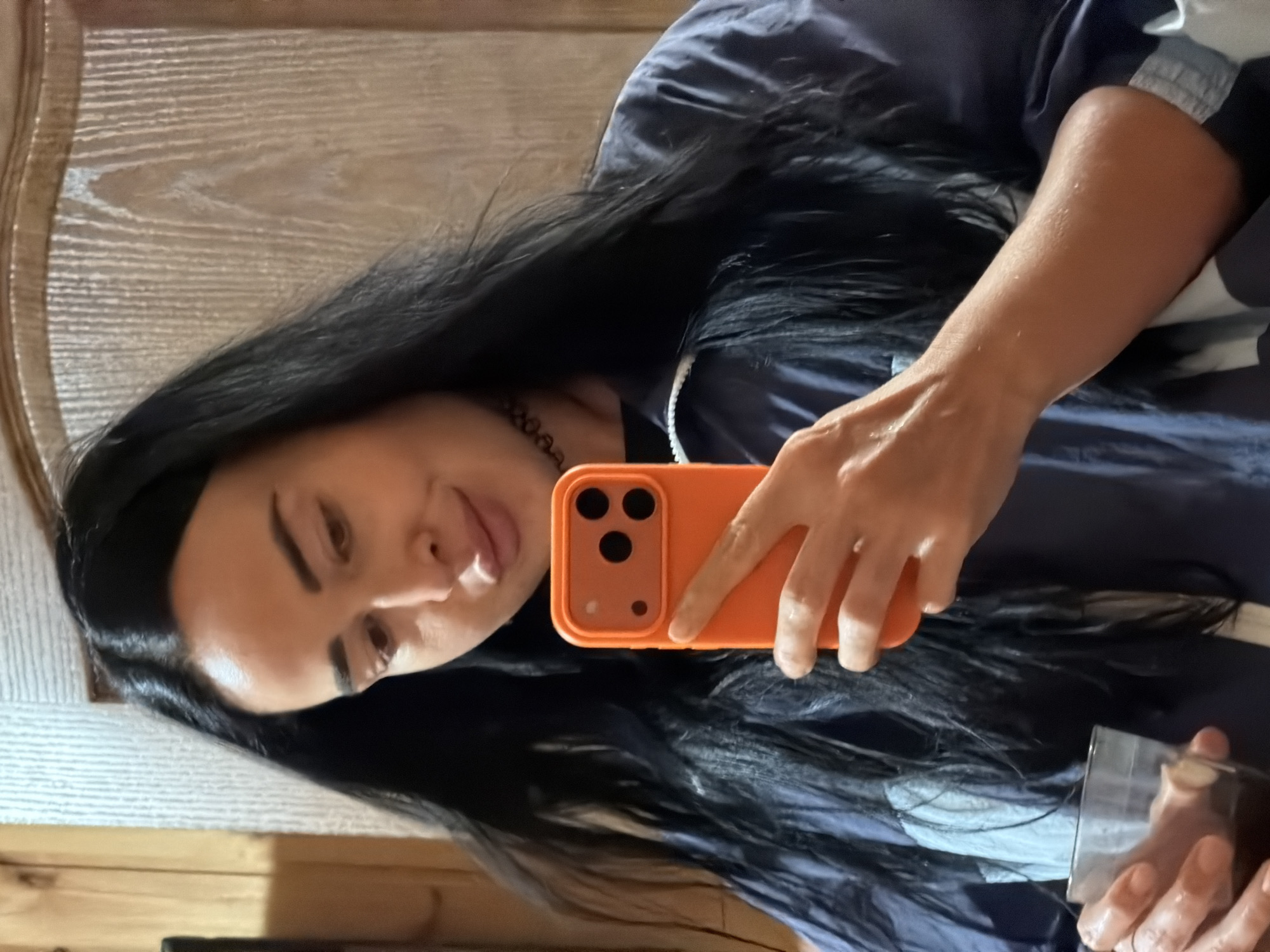 A woman with long, dark hair and light-colored skin is taking a selfie in a room. She is wearing a dark blue jacket with white accents and a black choker necklace. In her right hand, she holds an orange smartphone, and in her left hand, she holds a clear glass with a dark liquid. The background appears to be a wall with wooden paneling and textured white sections, possibly indicative of an interior setting in Sochi, Russia.