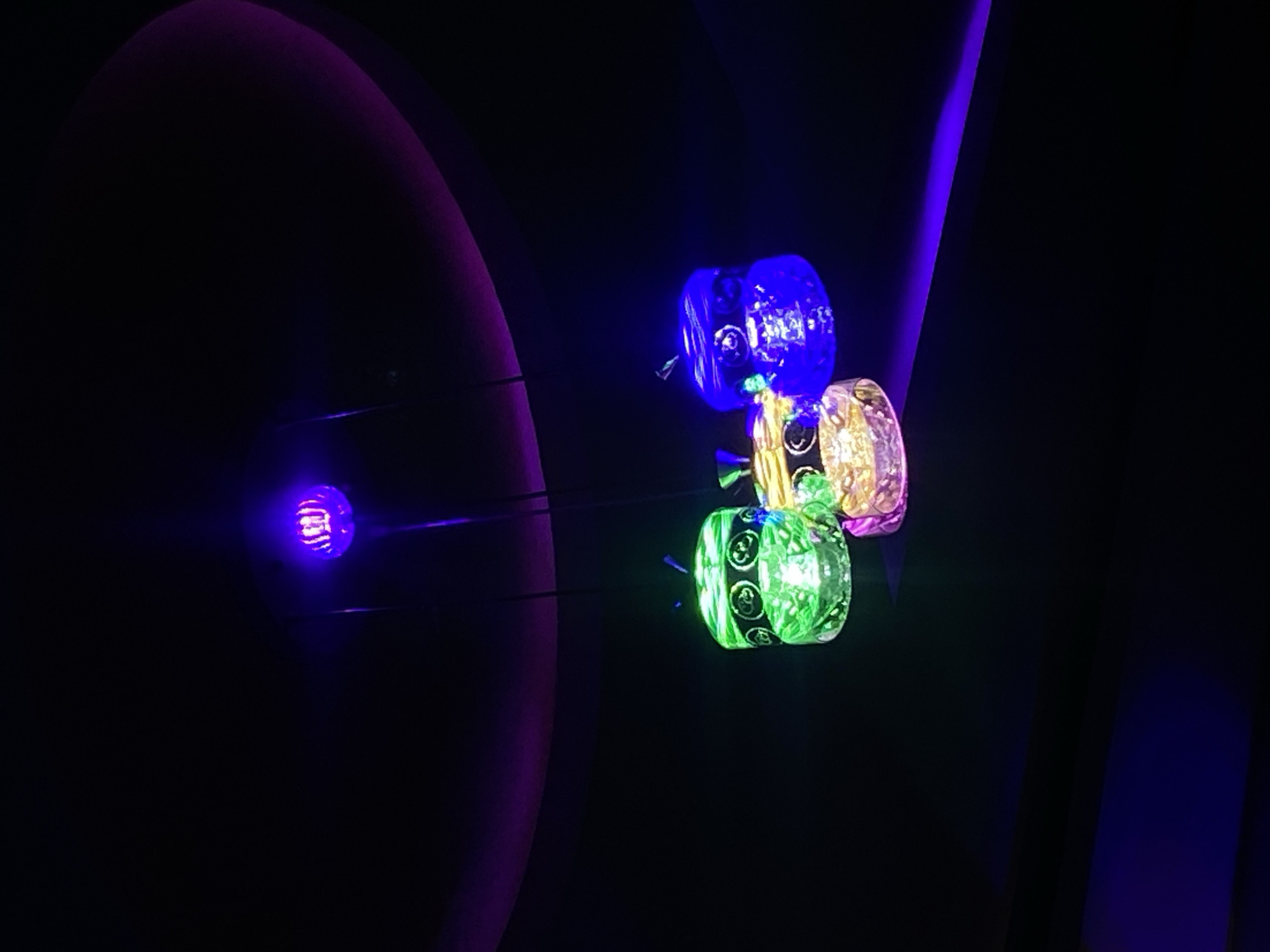 The image shows a dark setting with illuminated objects. At the left side of the image, there is a purplish, circular light. To the right of this, there are three small, stacked, cylinder-shaped items that are illuminated in blue, pink, and green, respectively. The cylinder-shaped items appear to have smiley face symbols on them. A beam of blue light is visible on the right side of the image. The city of Vom, Nigeria is not evident in this image.