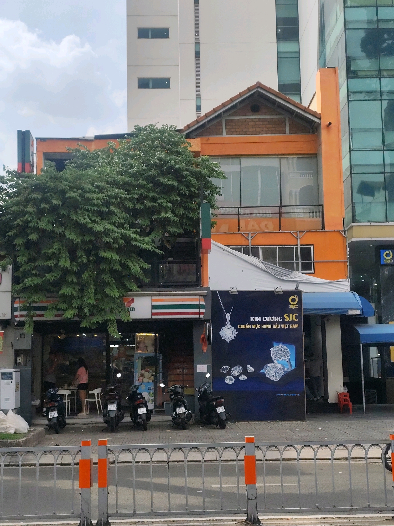 This image captures a street scene in Ho Chi Minh City, Vietnam, likely a commercial area. In the foreground, a row of parked motorcycles is visible on the sidewalk. Behind them, the facade of a building features a large advertisement for SJC jewelry, showcasing diamonds and the text "KIM CƯƠNG SJC CHUẨN MỰC HÀNG ĐẦU VIỆT NAM". Adjacent to this is a 7-Eleven store with its distinctive signage.
The architecture is a mix of modern and older structures. A tall, white building with multiple floors and rectangular windows stands prominently, partially obscured by an orange-painted building with a pitched roof. A large, leafy tree is positioned in front of the 7-Eleven, providing shade. Further to the right, a modern glass-facade building suggests a more contemporary commercial development. The street is paved, and a metal railing with orange and white bollards separates the sidewalk from the road. The sky is partly cloudy. Two individuals are visible inside the 7-Eleven store.