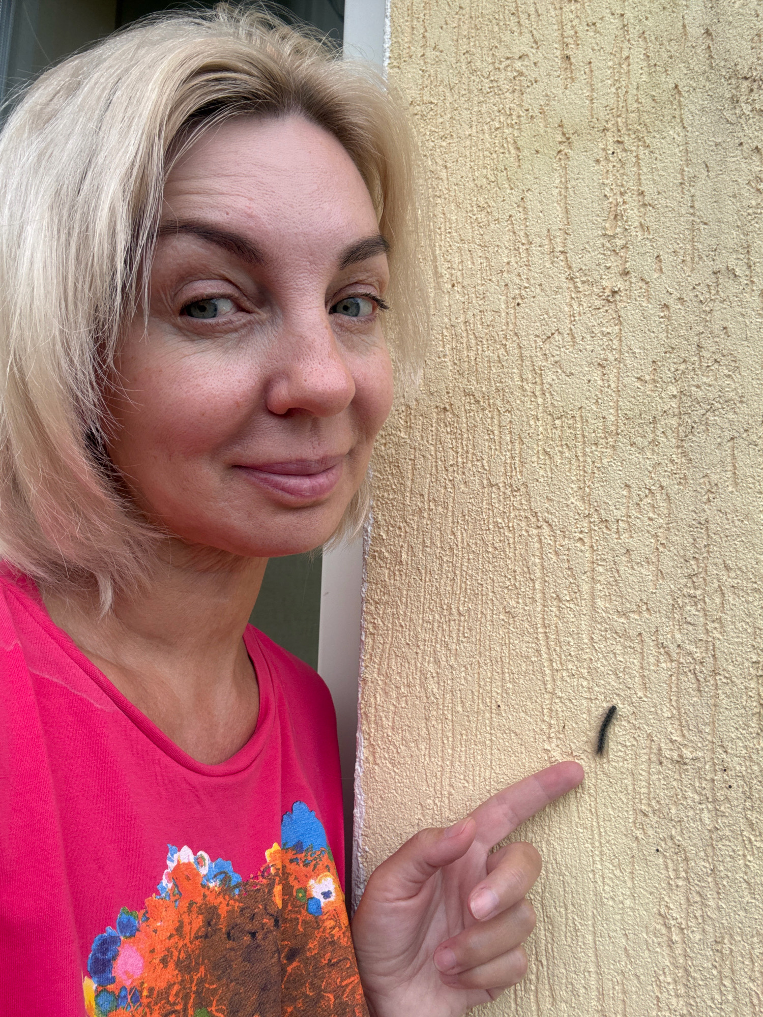 In Sochi, Russia, a woman with blonde hair and light-colored eyes, wearing a bright pink t-shirt with a colorful abstract design, is photographed from the chest up. She is standing next to a textured, light-yellow wall. Her left hand is visible, with her index finger extended towards a small, dark, segmented insect on the wall. The woman is looking at the camera and smiling slightly. The scene appears to be outdoors, with a hint of a green surface visible behind her to the left.