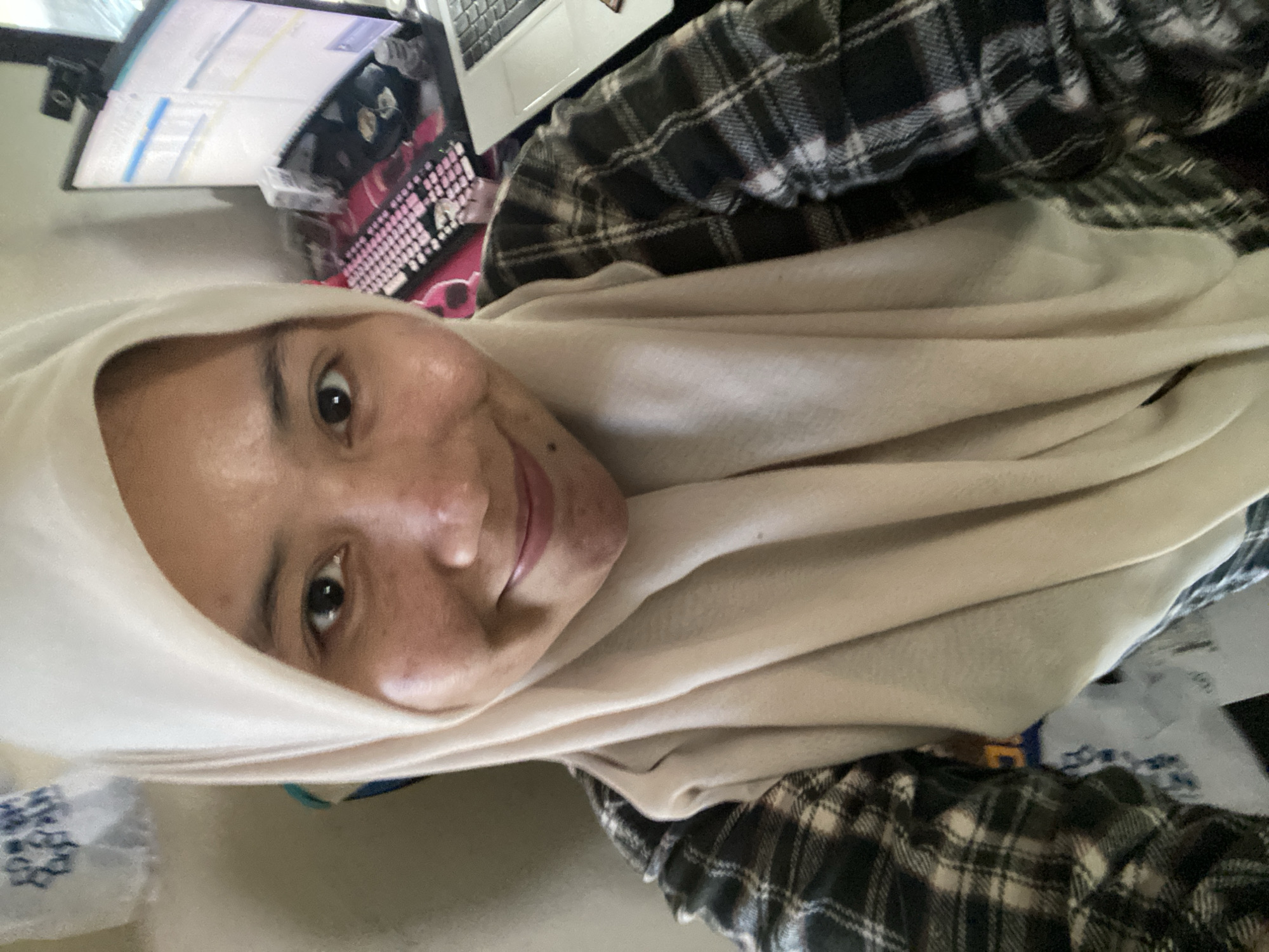A head-and-shoulders selfie of a woman wearing a light-colored hijab and a plaid shirt is shown from a close-up, slightly upward angle. The woman has fair skin, dark eyes, and is smiling faintly. Behind her, a computer monitor, keyboard, and laptop are visible, suggesting an indoor setting, possibly a home office or a workstation. A small web camera is mounted on top of the monitor. The background includes a wall and what appears to be some papers or bags. The image has a slightly grainy quality, typical of a smartphone camera.