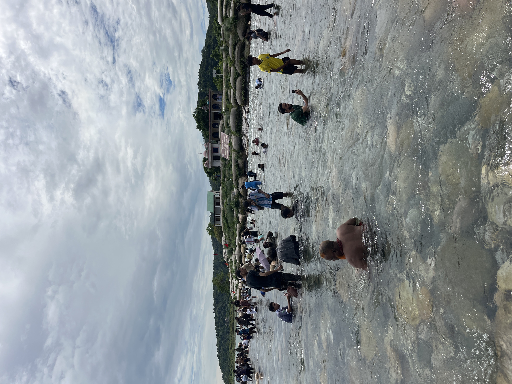 In Majai, India, a group of people are wading in a shallow, rocky river. The water is clear enough to see the riverbed, which is covered in stones of various sizes. The people are of different ages and genders, dressed in casual clothing, and some are partially submerged in the water. There are scattered structures visible on the opposite bank, including buildings and what appear to be religious sites, possibly temples or shrines, with domed roofs and colorful facades. Hills covered in lush green vegetation form the background. The sky above is cloudy, with patches of blue visible, suggesting a mix of sun and shade. Some individuals are engaged in activities such as taking photos with their phones, holding umbrellas, or simply standing and observing. The overall impression is one of a popular gathering spot for recreational activities or religious rituals.