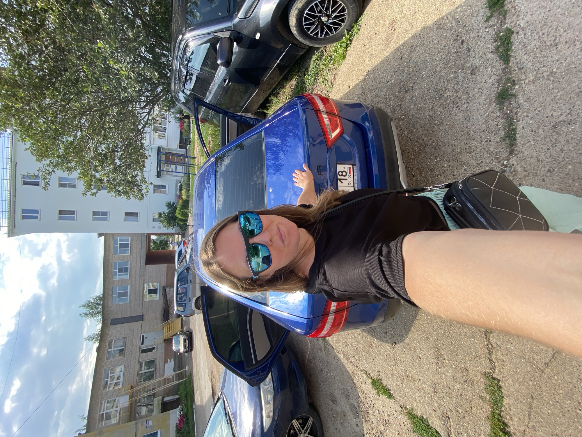 A woman stands in front of a blue car in what appears to be an urban environment in Noviy, Russia. She is wearing a black top and sunglasses and has a handbag slung over her shoulder. Her left hand is resting on the trunk of the blue car. Behind the blue car, a dark colored SUV is parked. In the background, there are multi-story buildings, one of which is light blue and another is made of brick. There are also trees and a cloudy sky. The ground is a paved surface with some patches of grass.